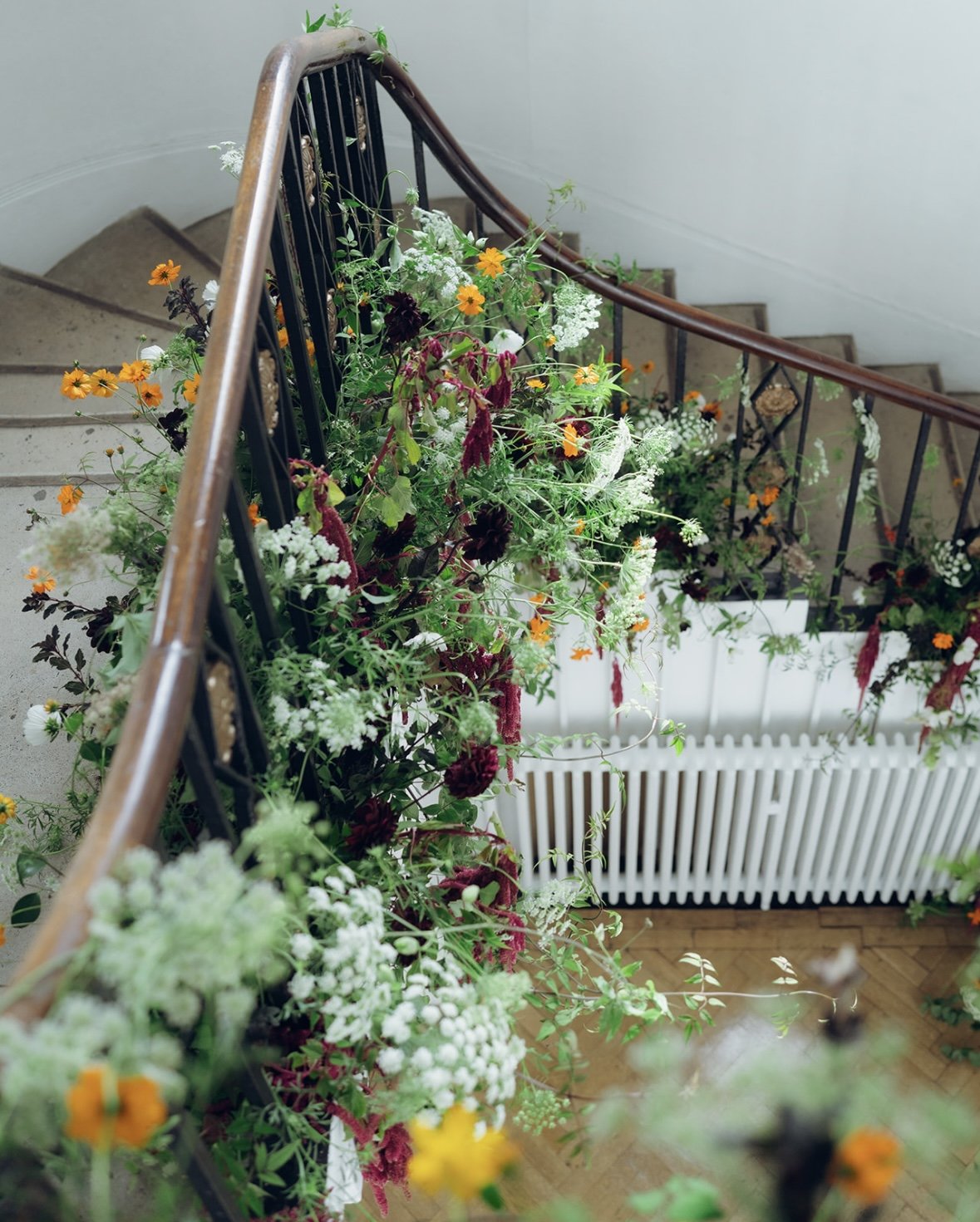 Indoor staircase with a black metal railing, decorated with colorful flowers and green plants, next to a white radiator under a light-colored wall.