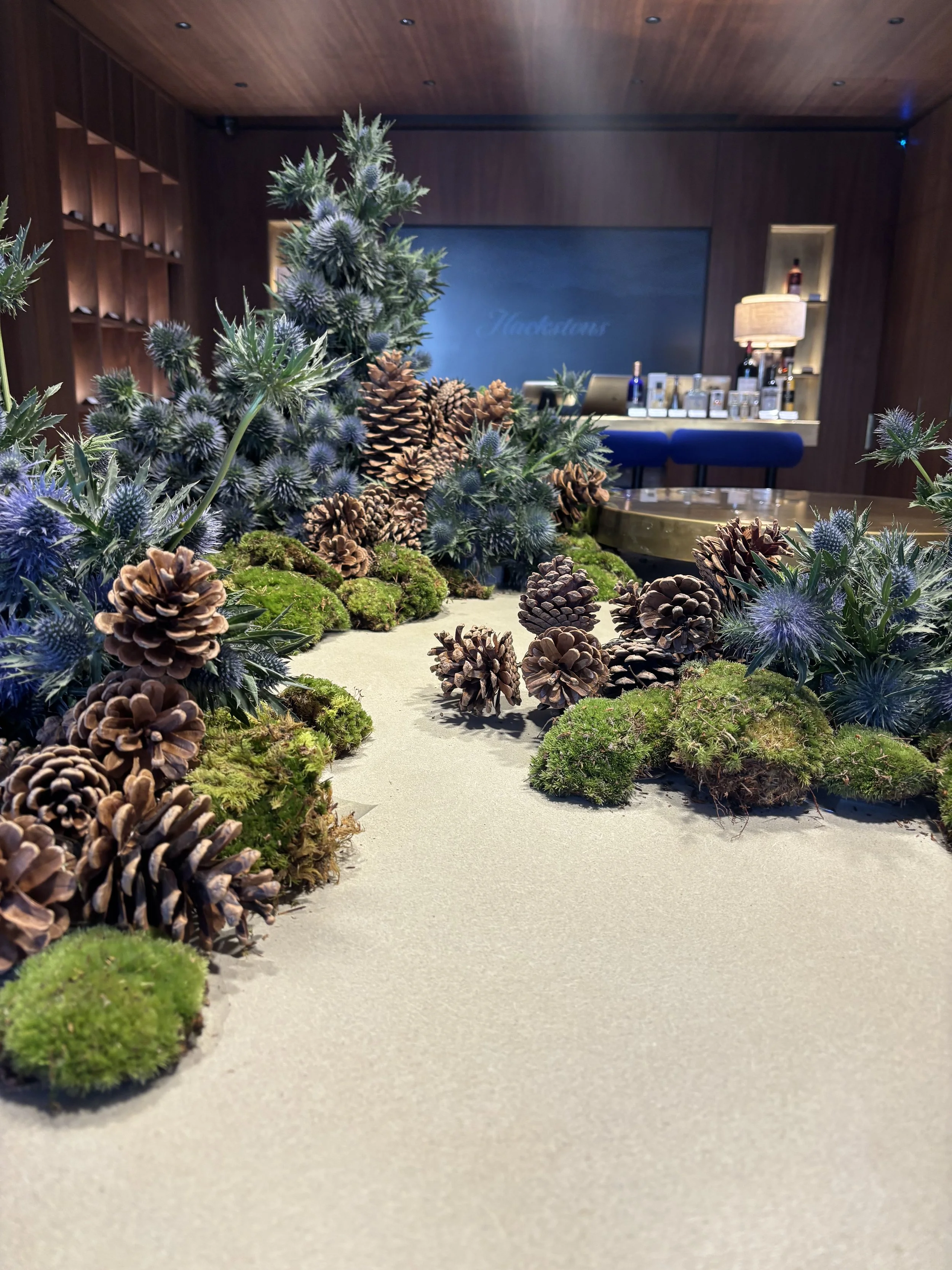 Decorative indoor scene with pinecones, moss, and artificial plants arranged on a light-colored surface, with a dark wood-paneled room and bar area in the background.