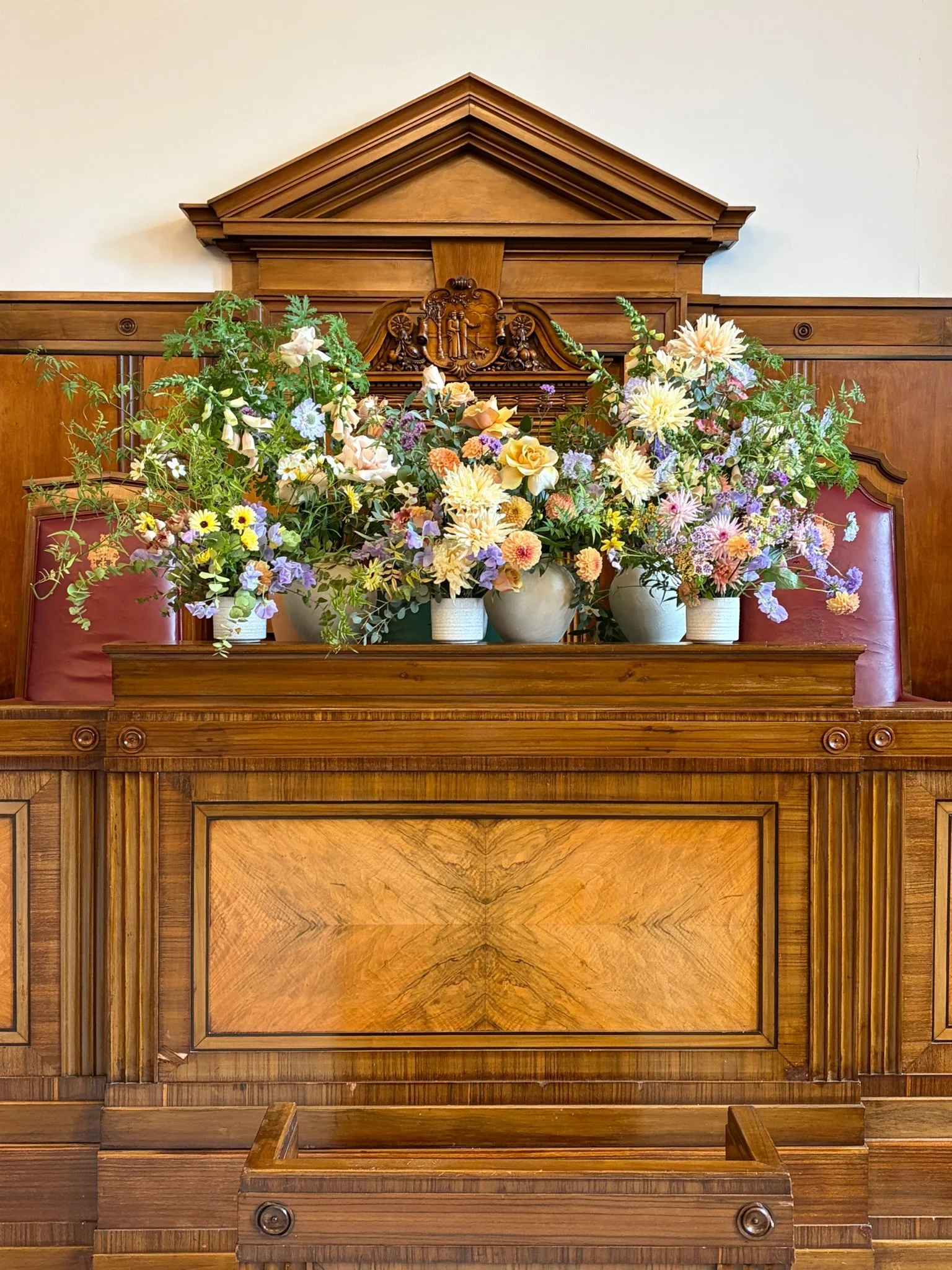 A wooden judge's bench with three flower arrangements in vases on top, set against a wood panel backdrop in a court or ceremonial room.