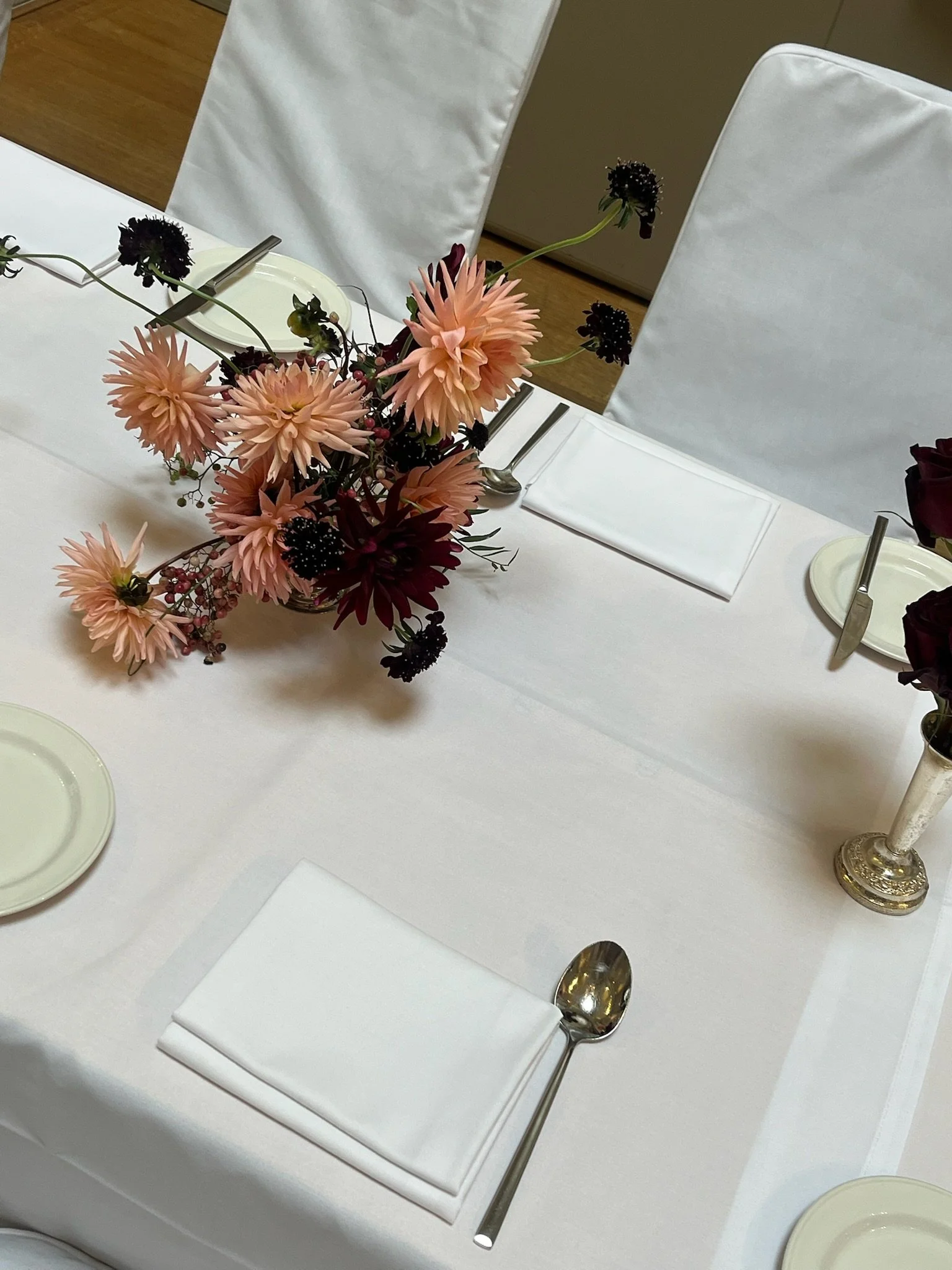 A table decorated with a floral centerpiece featuring pink and dark purple flowers, set with white plates, silverware, and white napkins, and surrounded by white chairs.