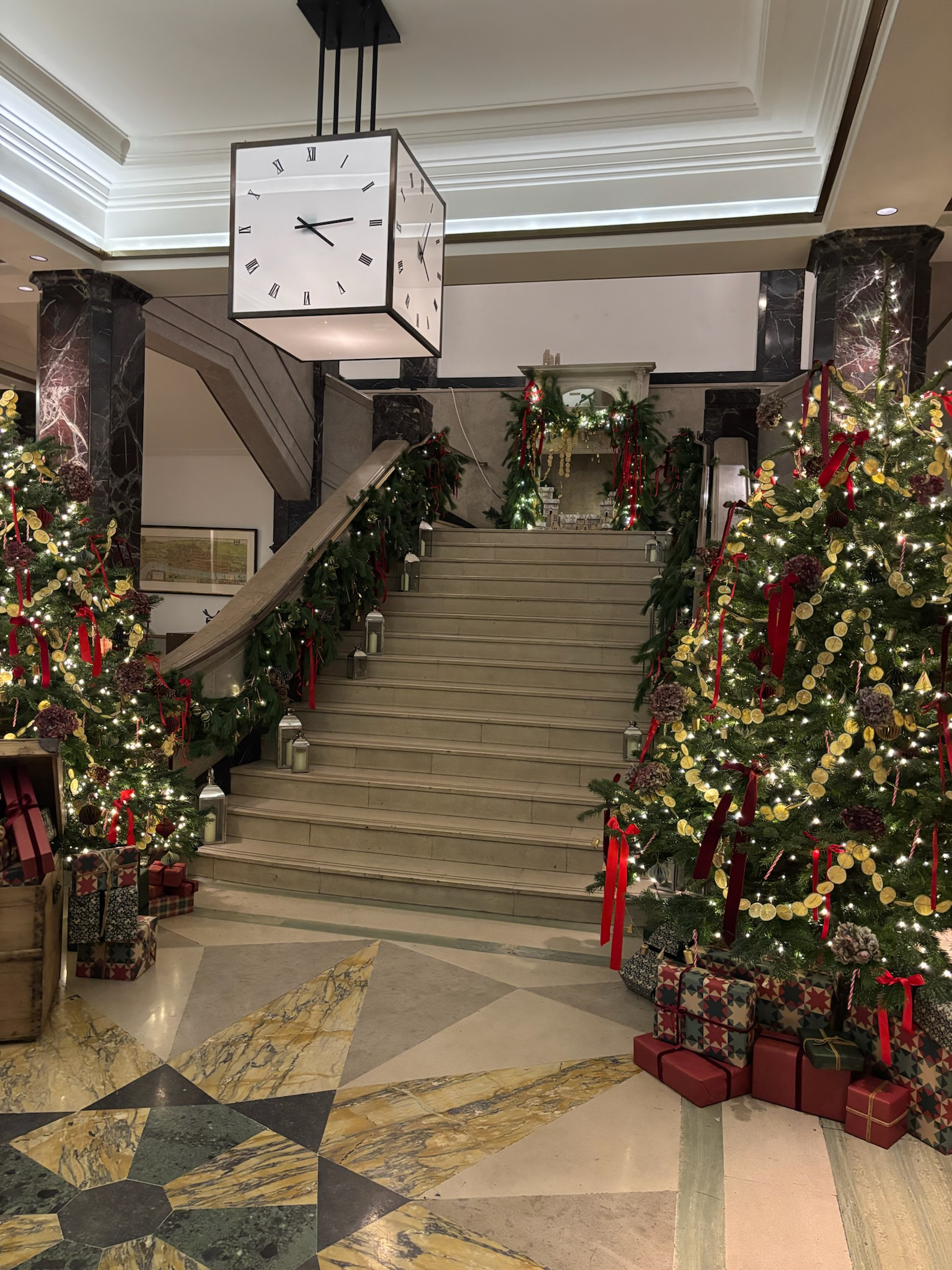 Decorated Christmas trees and wrapped presents on the floor beside a staircase in a hotel lobby with Christmas garlands and a large ceiling clock.