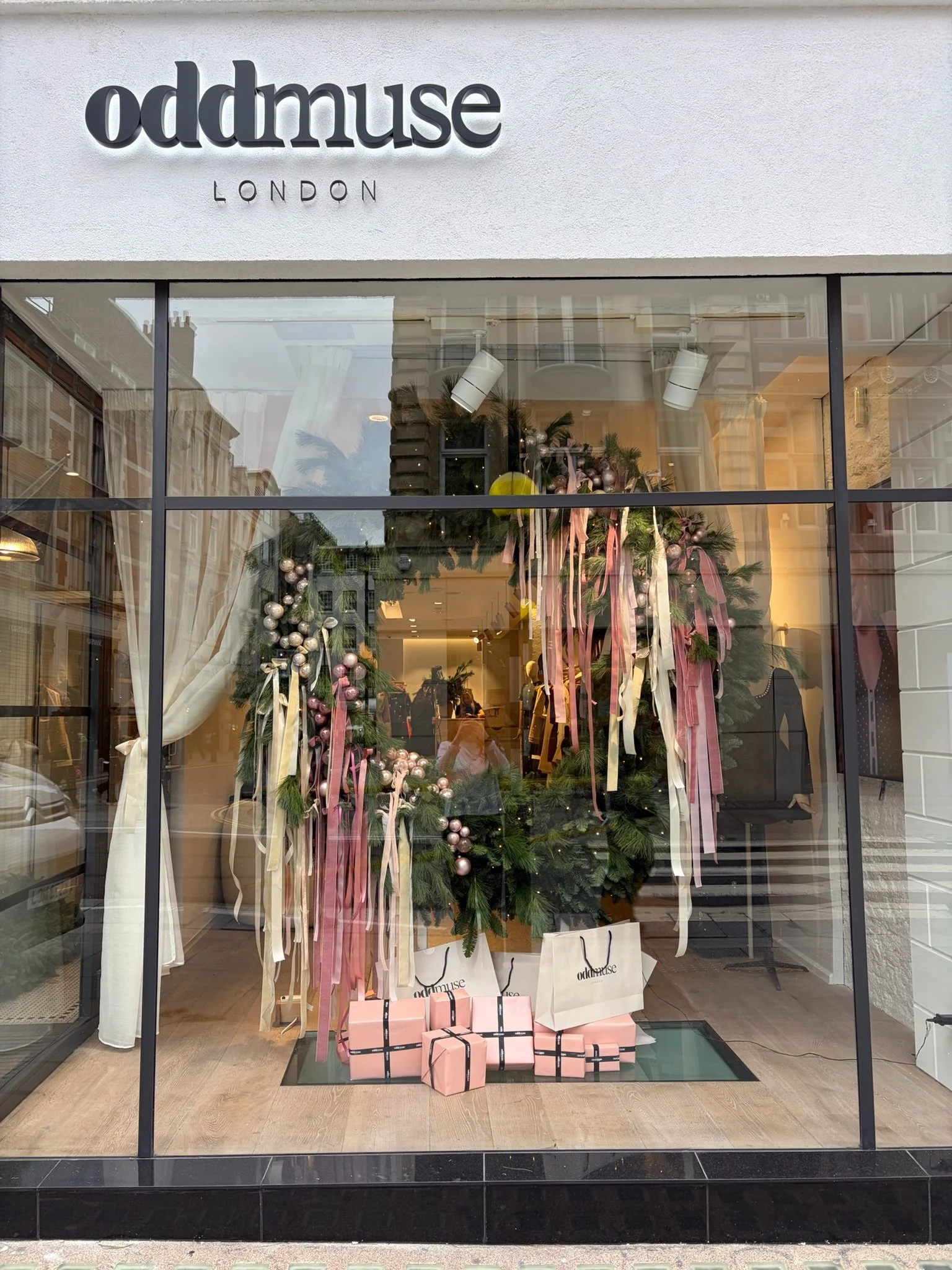 Store window display with Christmas decorations including a large wreath with pink ribbons, ornaments, and wrapped gift boxes, with the store name 'oddmuse LONDON' above the window.