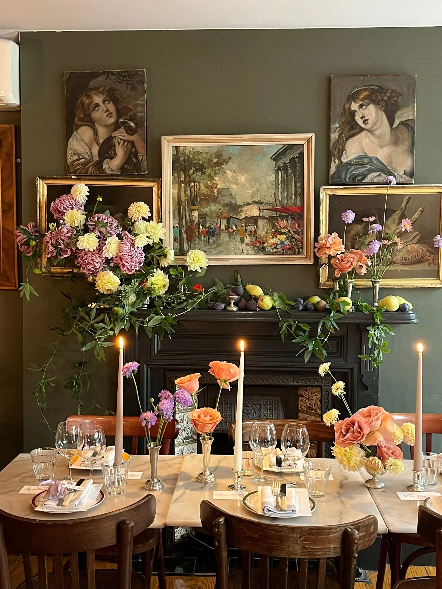 A beautifully set dining table with floral centerpieces, tall pink and white candles, and elegant glassware. The background features a green wall adorned with framed paintings and a colorful impressionist cityscape, with a decorative mantle holding more flowers and fruit.