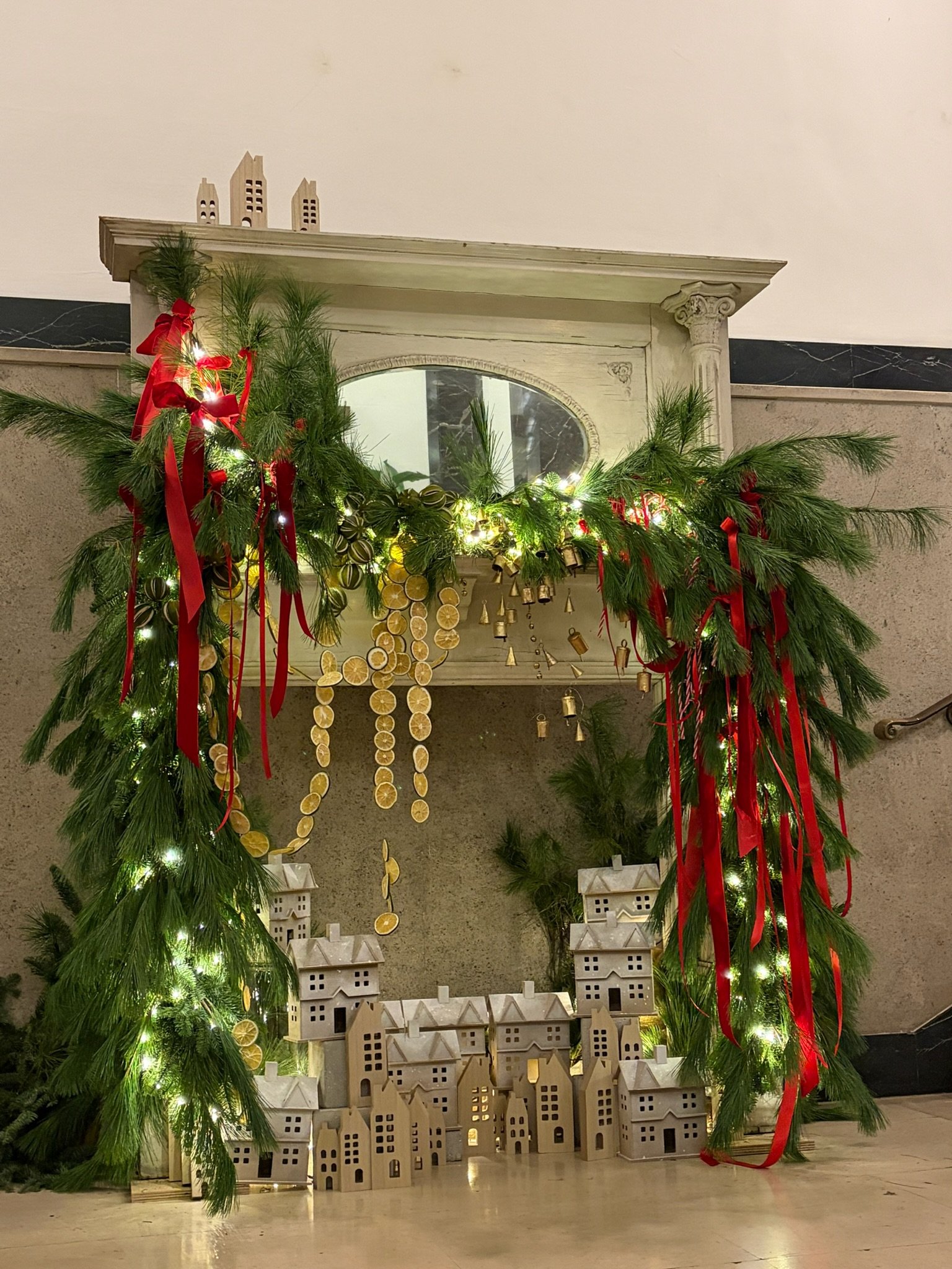 Holiday display with green pine garland decorated with red ribbons, gold bells, and string lights surrounding a fireplace. Golden paper ornaments with lemon slice designs hang from the garland. Small wooden house decorations are placed at the base of