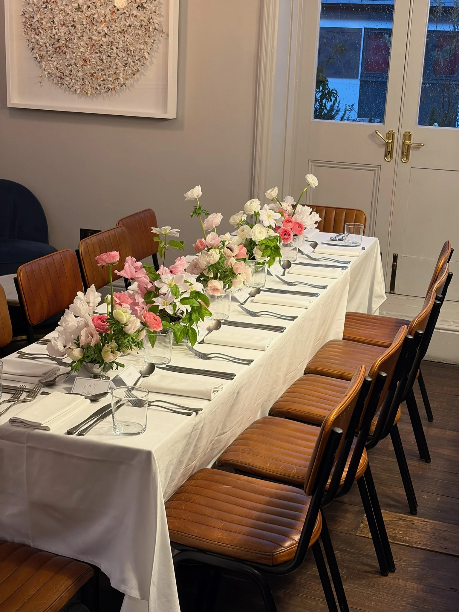 A long dining table set with white tablecloth, glassware, silverware, white napkins, and pink and white floral centerpieces, in a well-lit room with a large abstract artwork on the wall and closed double doors in the background.