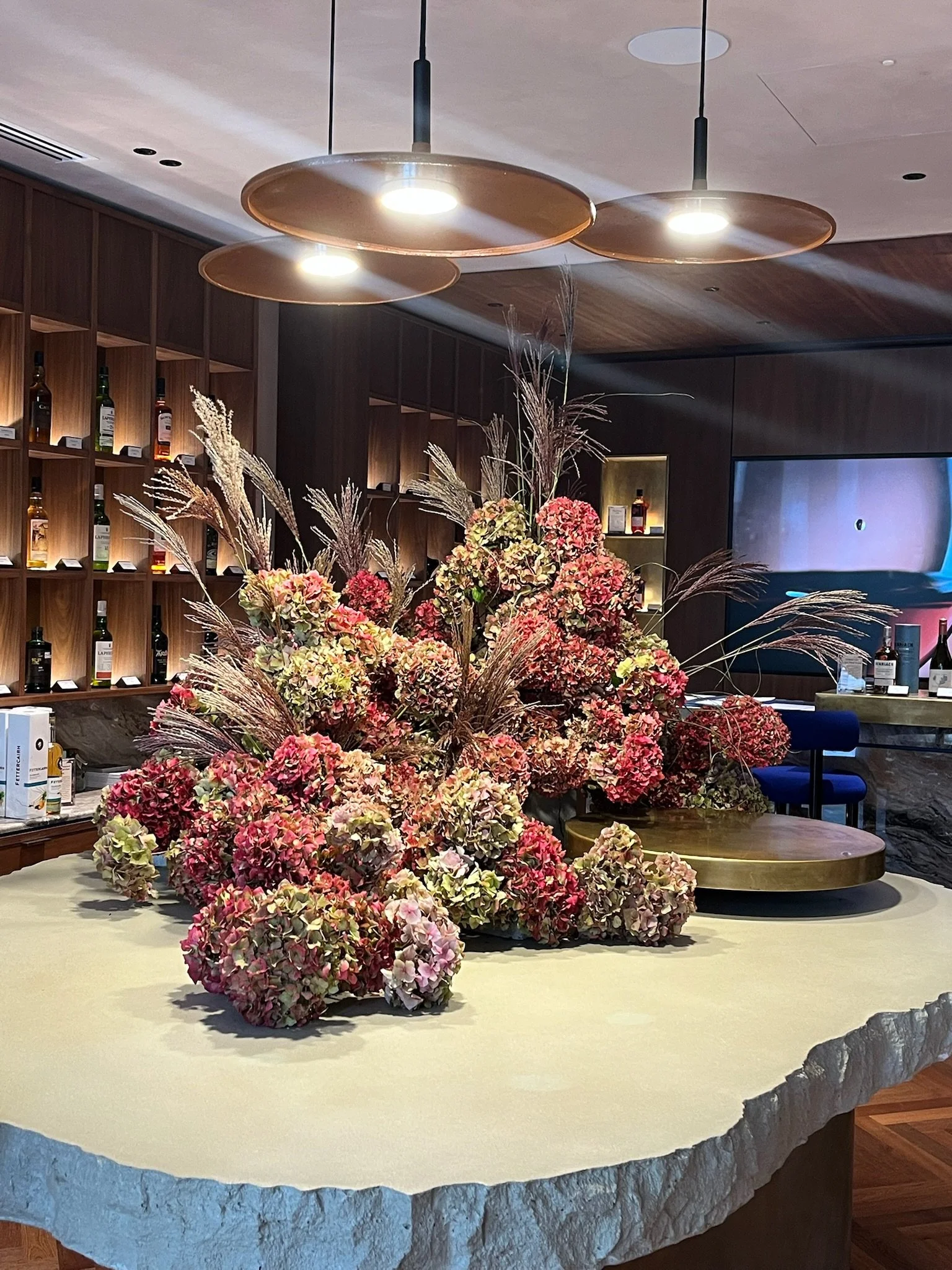 A large floral arrangement with pink and white hydrangeas and tall grasses on a stone countertop in a modern interior with wooden shelves and a television.