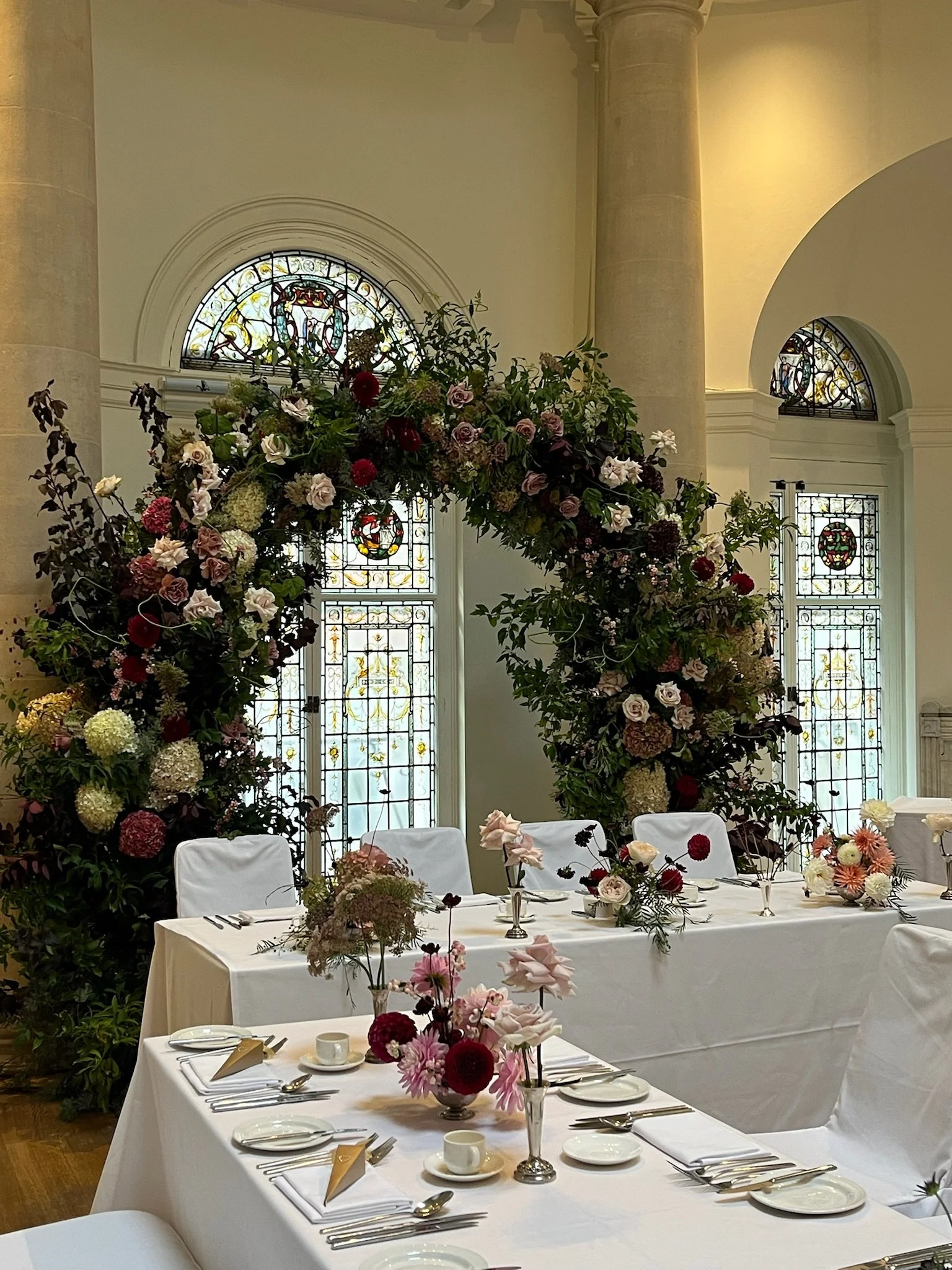 Wedding reception setup with white tablecloths, floral centerpieces, and a large floral arch in a decorated hall with stained glass windows.