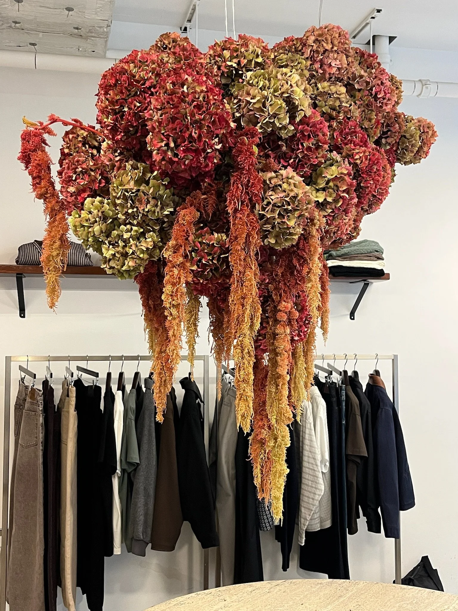 A large, colorful bouquet of dried hydrangea flowers and hanging amaranthus, suspended from the ceiling above a clothing rack filled with various garments in a retail setting.