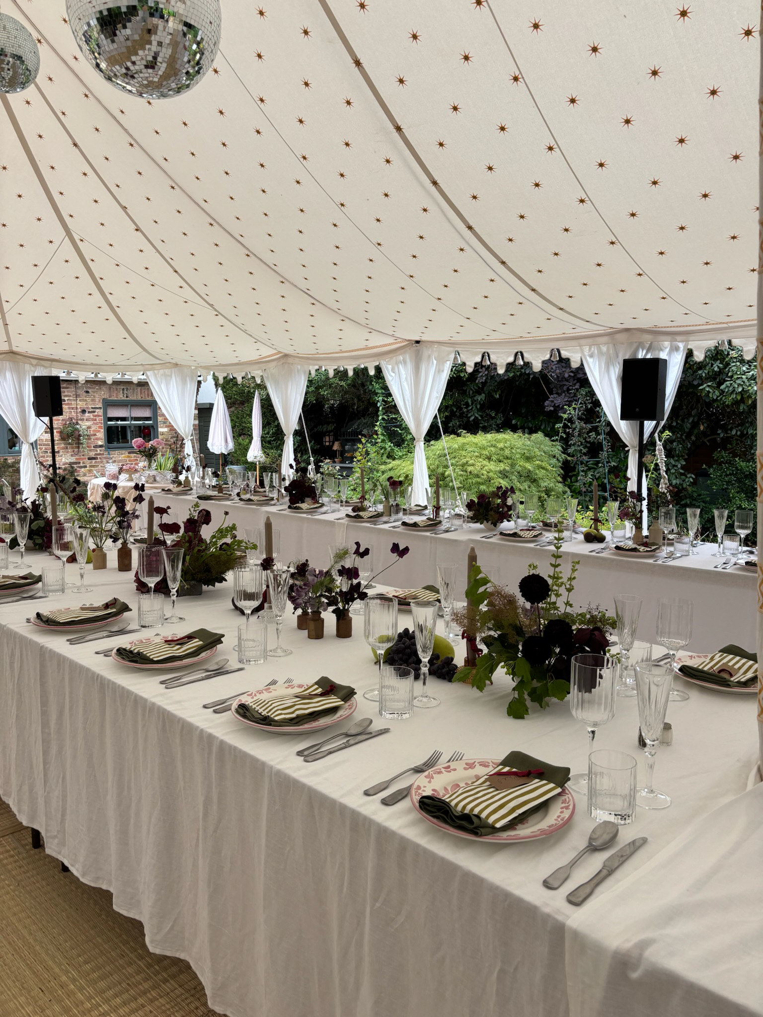 Elegant outdoor dining setup under a large tent with white draping, decorated with flowers, glassware, and tableware.