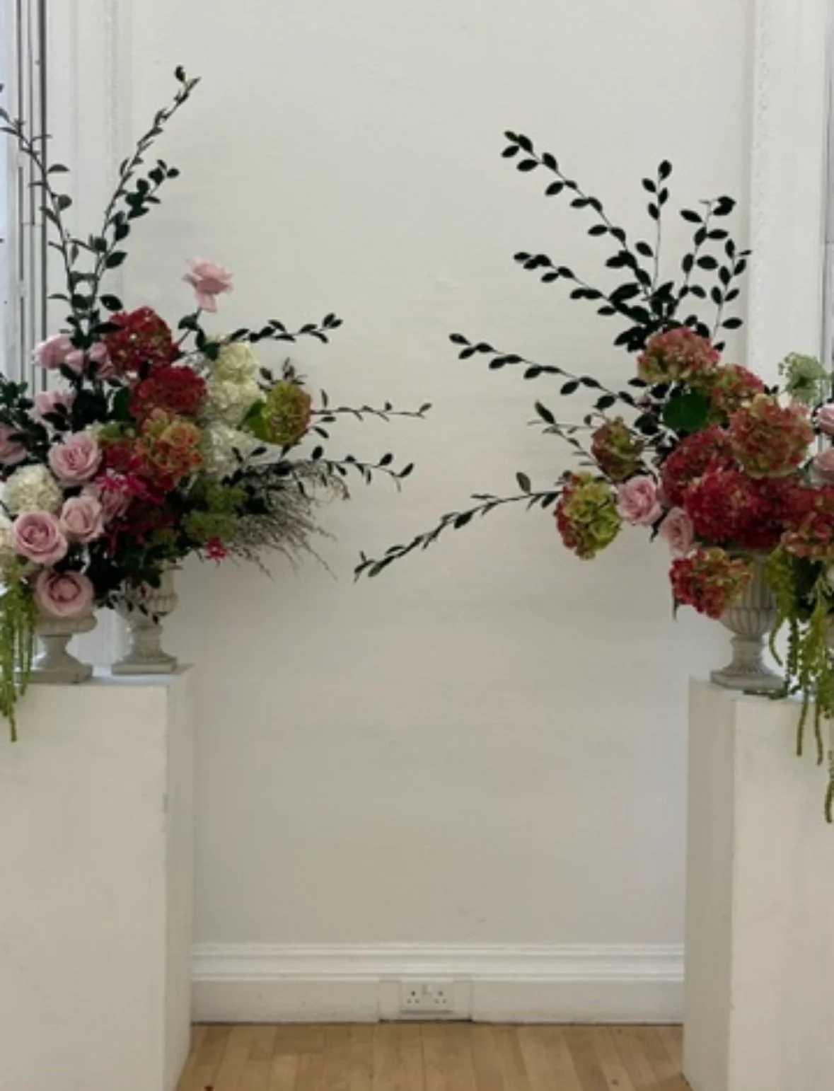 Two floral arrangements with pink and red flowers and dark green leaves on white pedestals against a cream-colored wall.