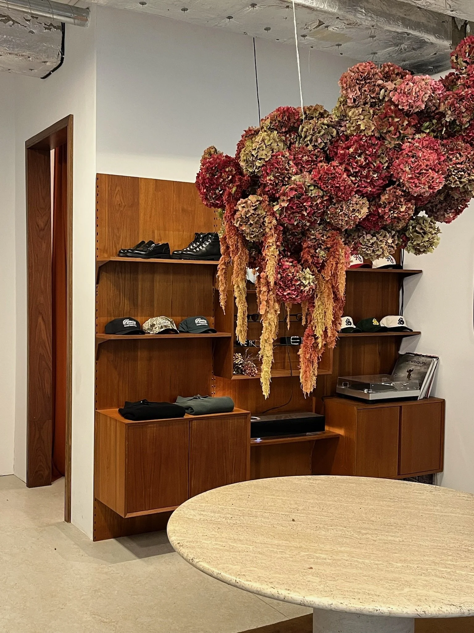A wooden cabinet with hats and shoes on its shelves and a turntable on top. Above, a large hanging arrangement of dried flowers in shades of pink, red, and green.