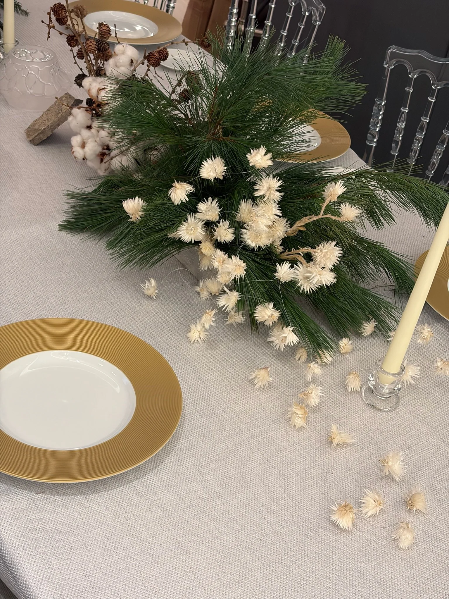 A table centerpiece with green pine branches, white flowers, and cotton stems, surrounded by gold-rimmed plates, a cream-colored candle, and a beige tablecloth.