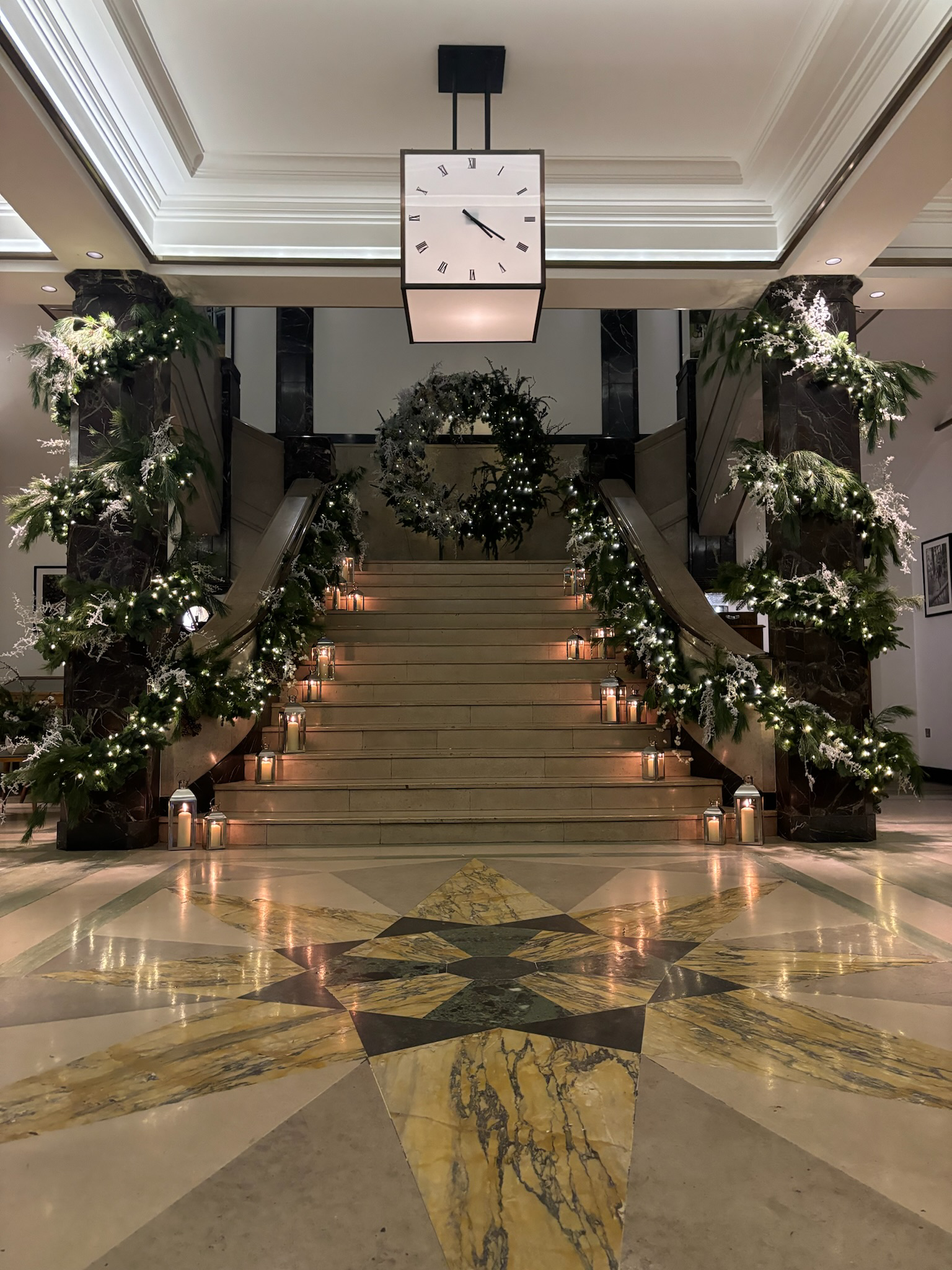 Decorated staircase with Christmas wreath and white string lights, surrounded by greenery and candles, inside a building with a large clock overhead.