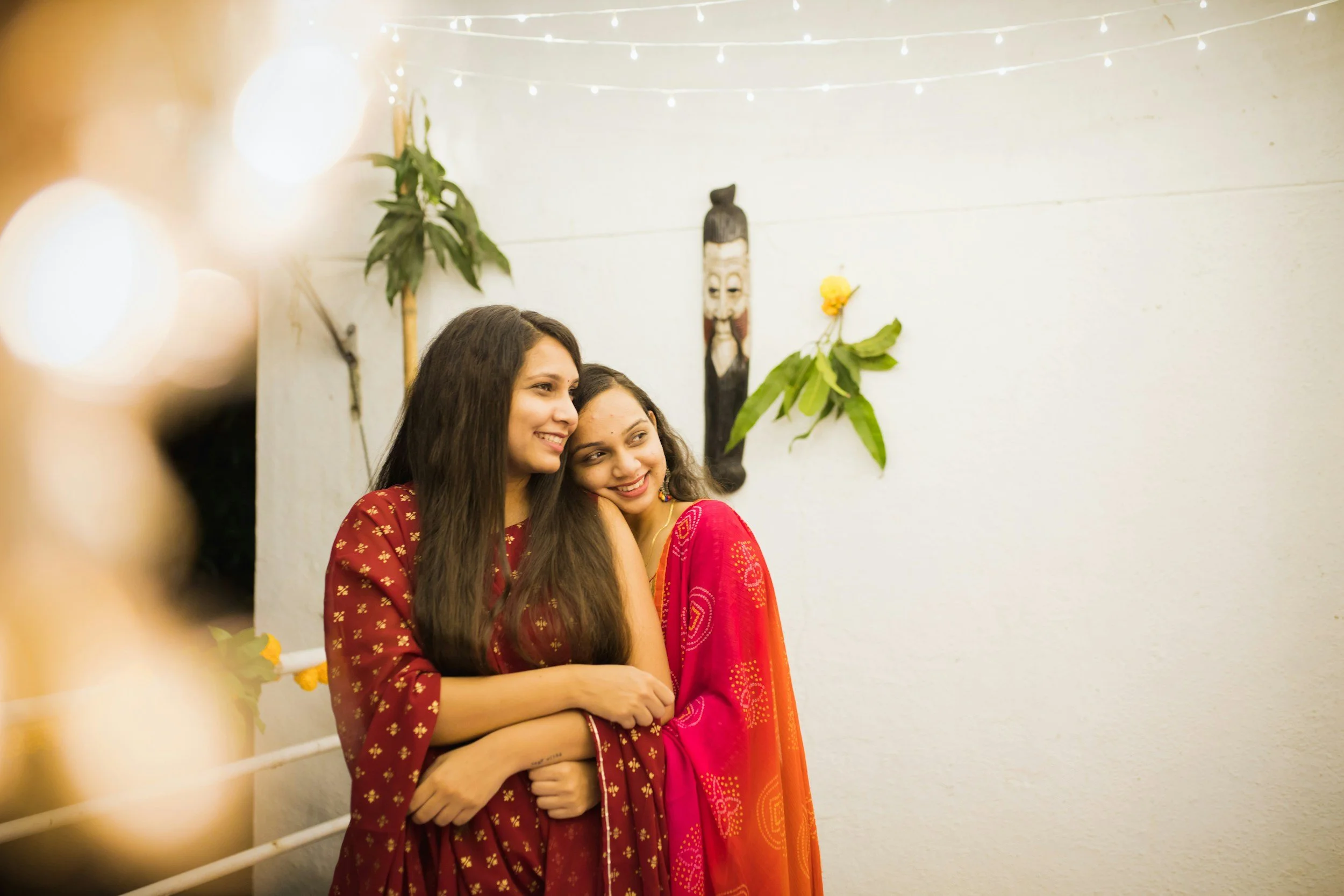 Two women, one in a red traditional dress with gold details and the other in a pink traditional dress, standing close together and smiling inside a decorated room with hanging lights, a wall hanging mask, and tropical leaves.