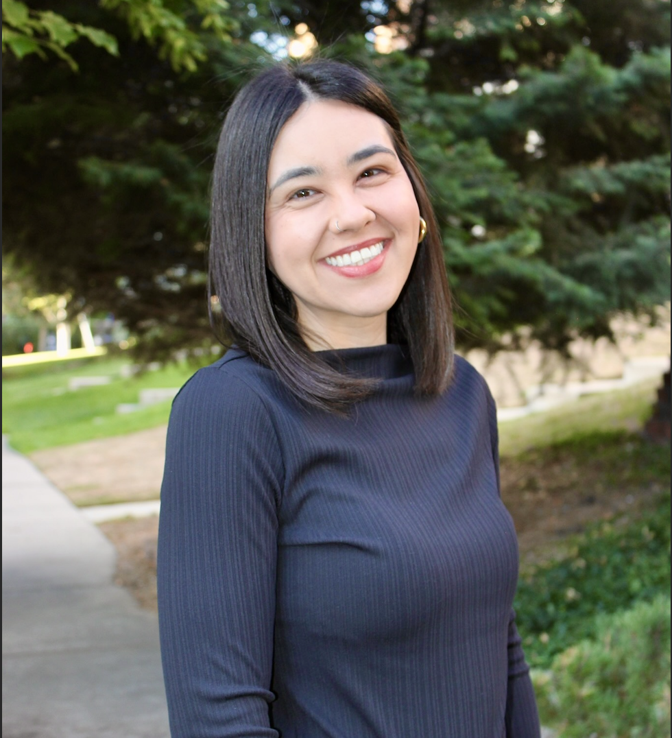 A young woman with shoulder-length dark hair, wearing a navy long-sleeve top, smiling outdoors with trees and a sidewalk in the background.
