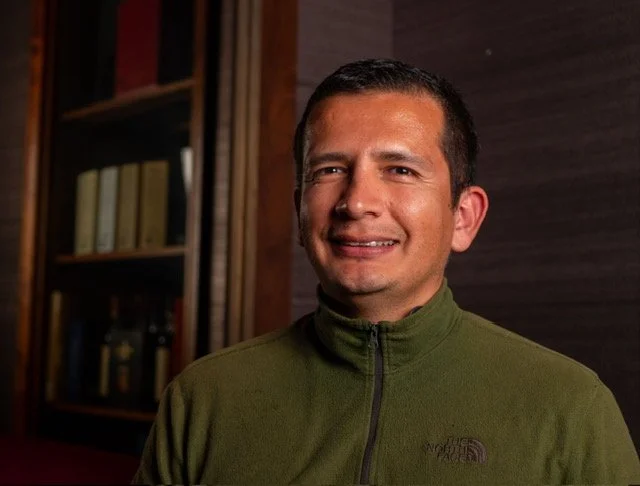 A man smiling, wearing a green North Face jacket, with a bookshelf in the background.