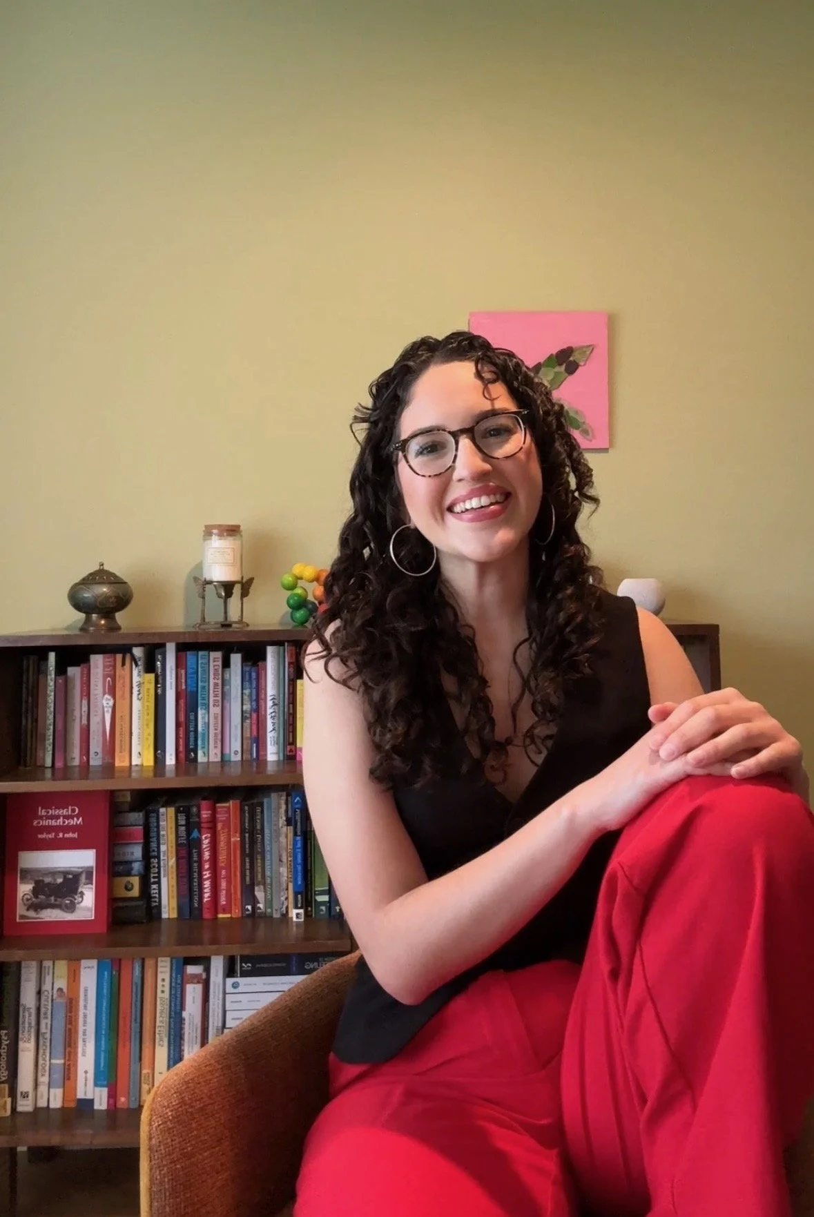 A woman with curly dark hair, wearing glasses, a black sleeveless top, and red pants, sitting on an orange chair in front of a bookshelf with books and decorative items, smiling at the camera.