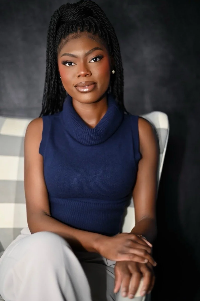 Portrait of a young African American woman with long braided hair, wearing a sleeveless navy blue turtleneck top, sitting against a dark background.