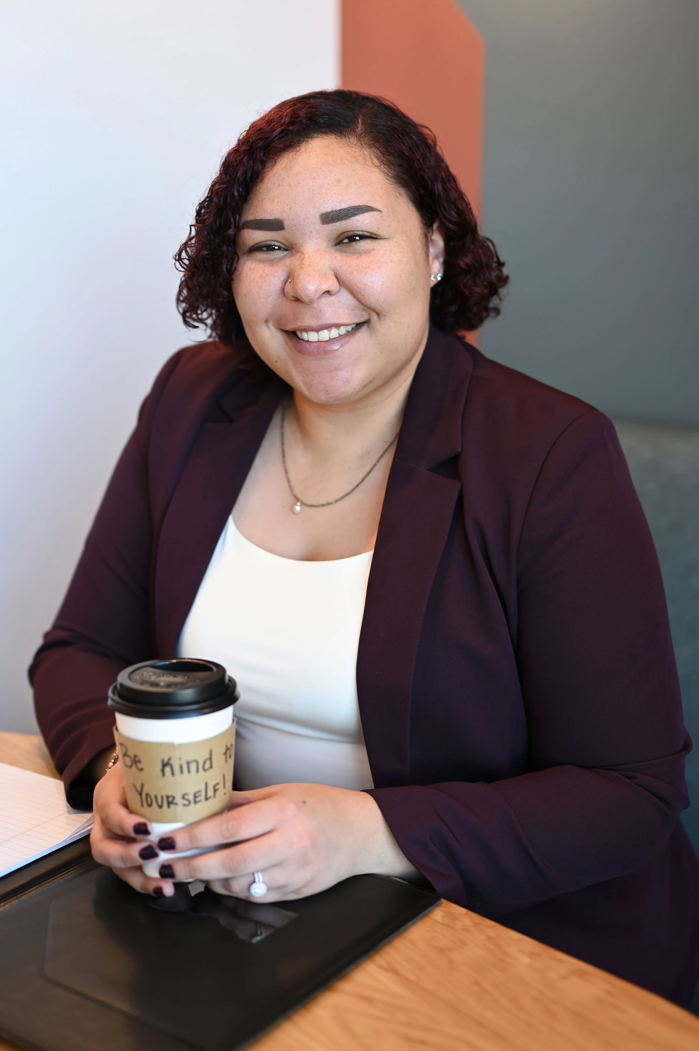 A woman with short brown curly hair, wearing a maroon blazer and white top, smiling while holding a coffee cup with a handwritten message that says, 'Be kind to YOURSELF!' She is sitting at a table with a laptop and notebook, in an indoor setting with a grey and peach color wall in the background.