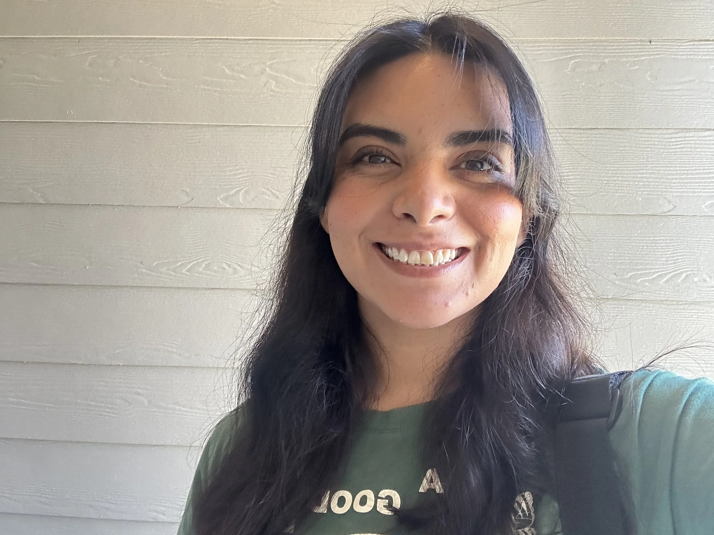 A woman with long dark hair smiling in front of a light-colored wooden wall, wearing a dark green shirt.