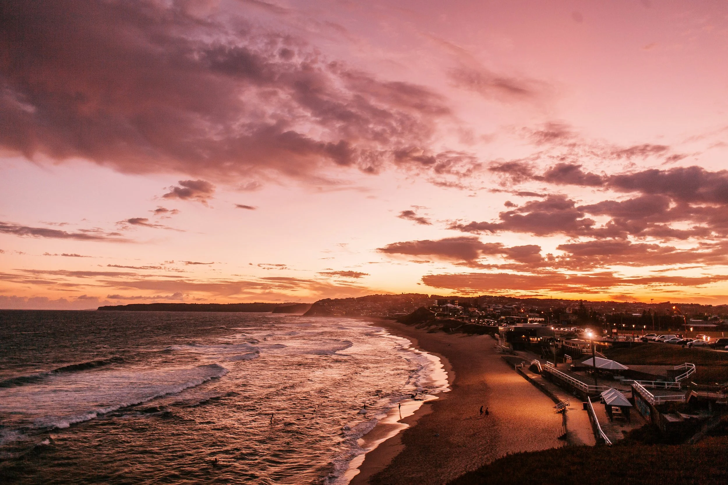 A sunset over a beach with a cloudy sky, ocean waves, and a shoreline with buildings and parking lot.