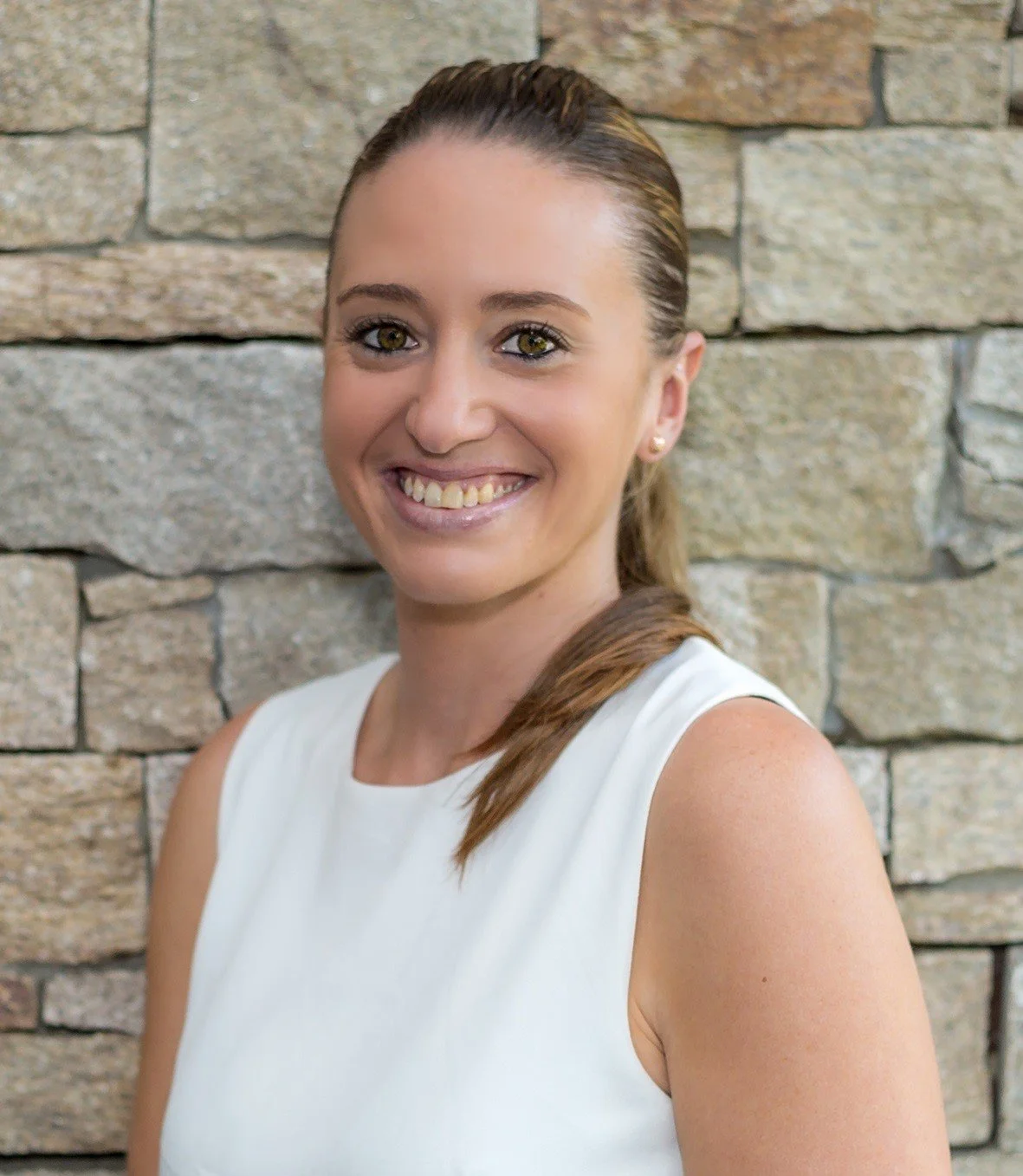 A smiling woman with brown hair in a ponytail, wearing a white sleeveless top, standing in front of a stone wall.
