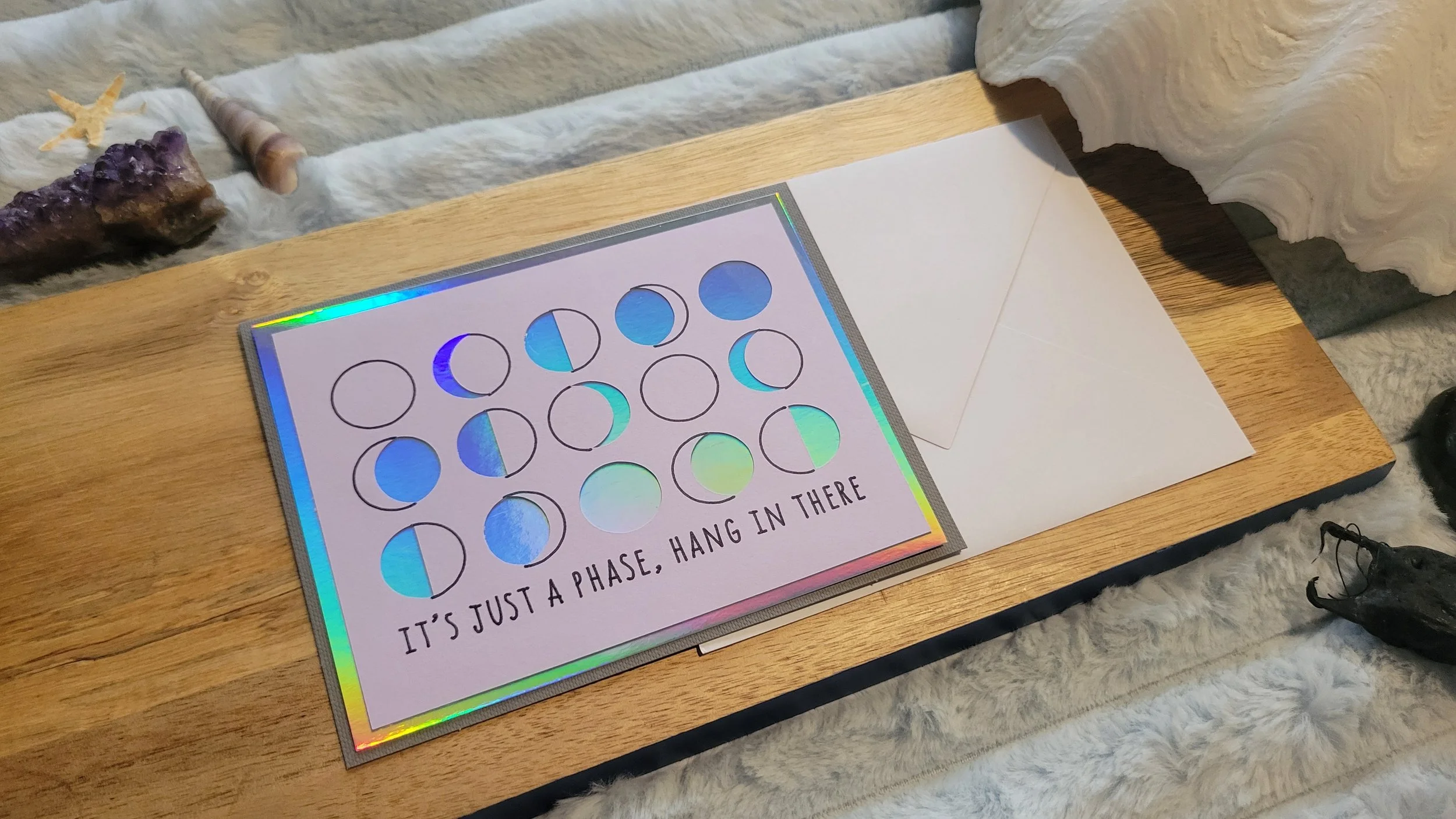 Handmade greeting card featuring moon phases in a grid with iridescent foil accents and the phrase “It’s just a phase, hang in there” on a holographic border.