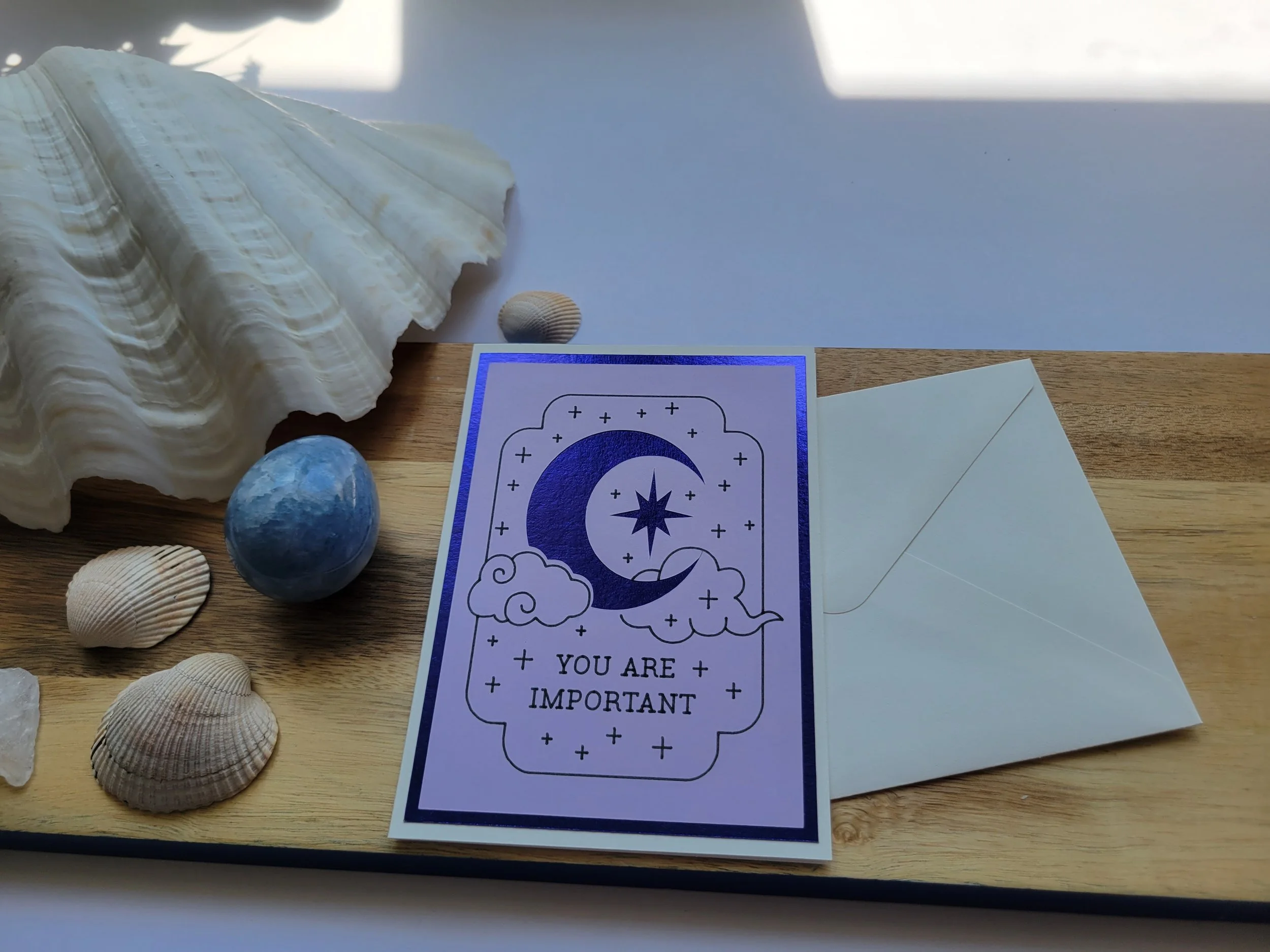 White Greeting card with blue metallic crescent moon and star design and the message “You Are Important,” placed on a wooden surface with seashells and a matching envelope.