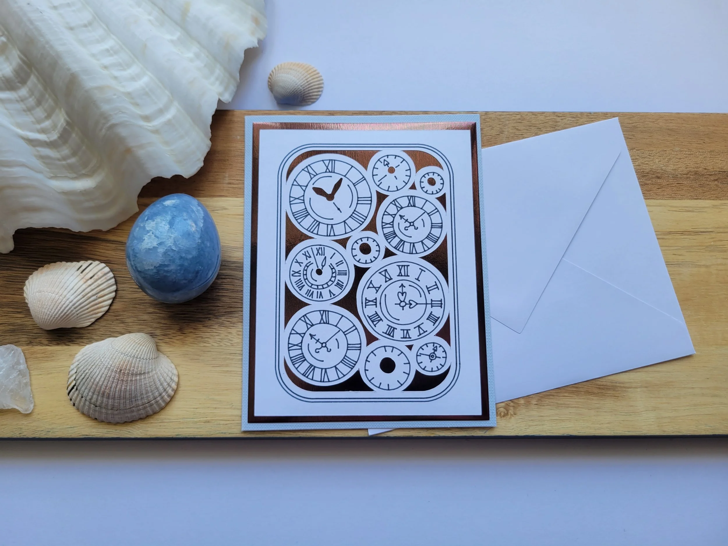 Close-up of a clock-themed greeting card with copper-toned foil accents and illustrated clock faces, displayed with a white envelope on a wooden board.