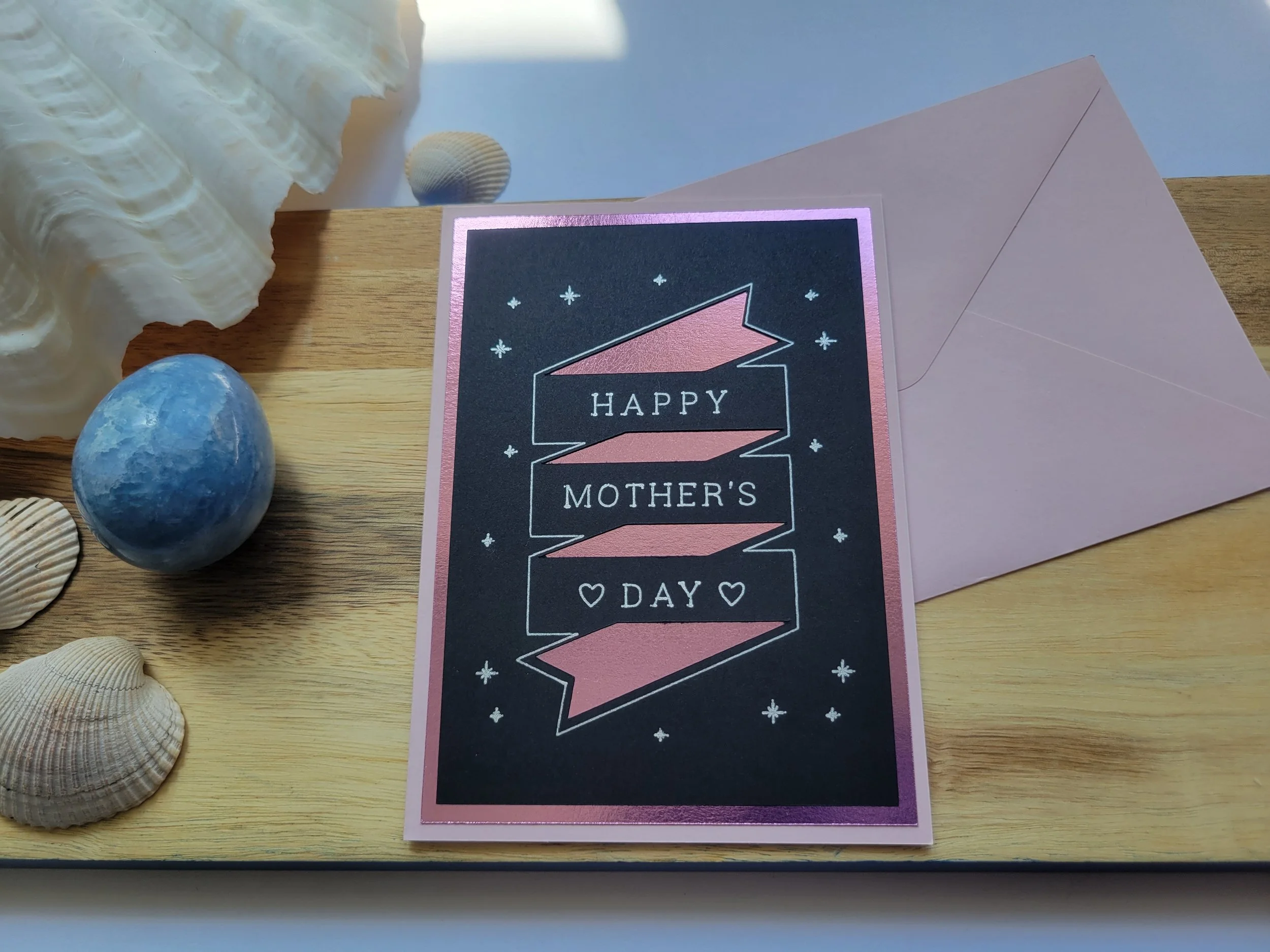 ink Greeting card with pink metallic banner design on black cardstock and the message “Happy Mother’s Day” placed on a wooden surface with seashells and a matching envelope.