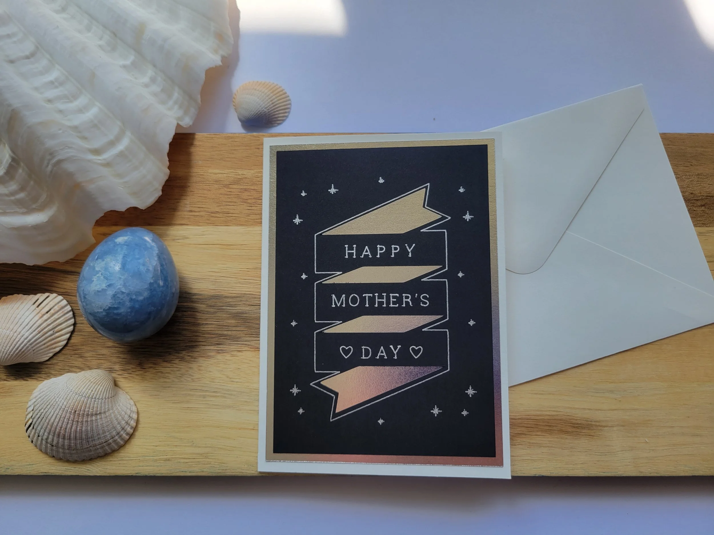 White Greeting card with silver metallic banner design on black cardstock and the message “Happy Mother’s Day” placed on a wooden surface with seashells and a matching envelope.