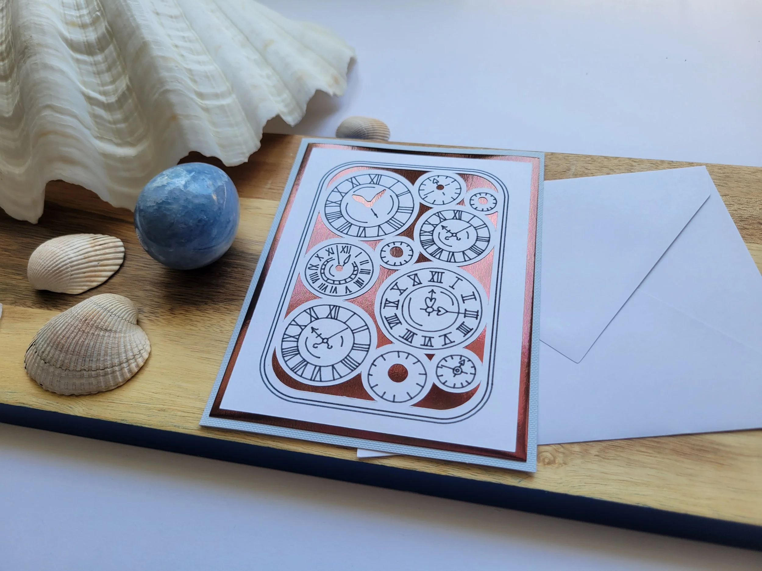 Close-up of a clock-themed greeting card with copper-toned foil accents and illustrated clock faces, displayed with a white envelope on a wooden board.