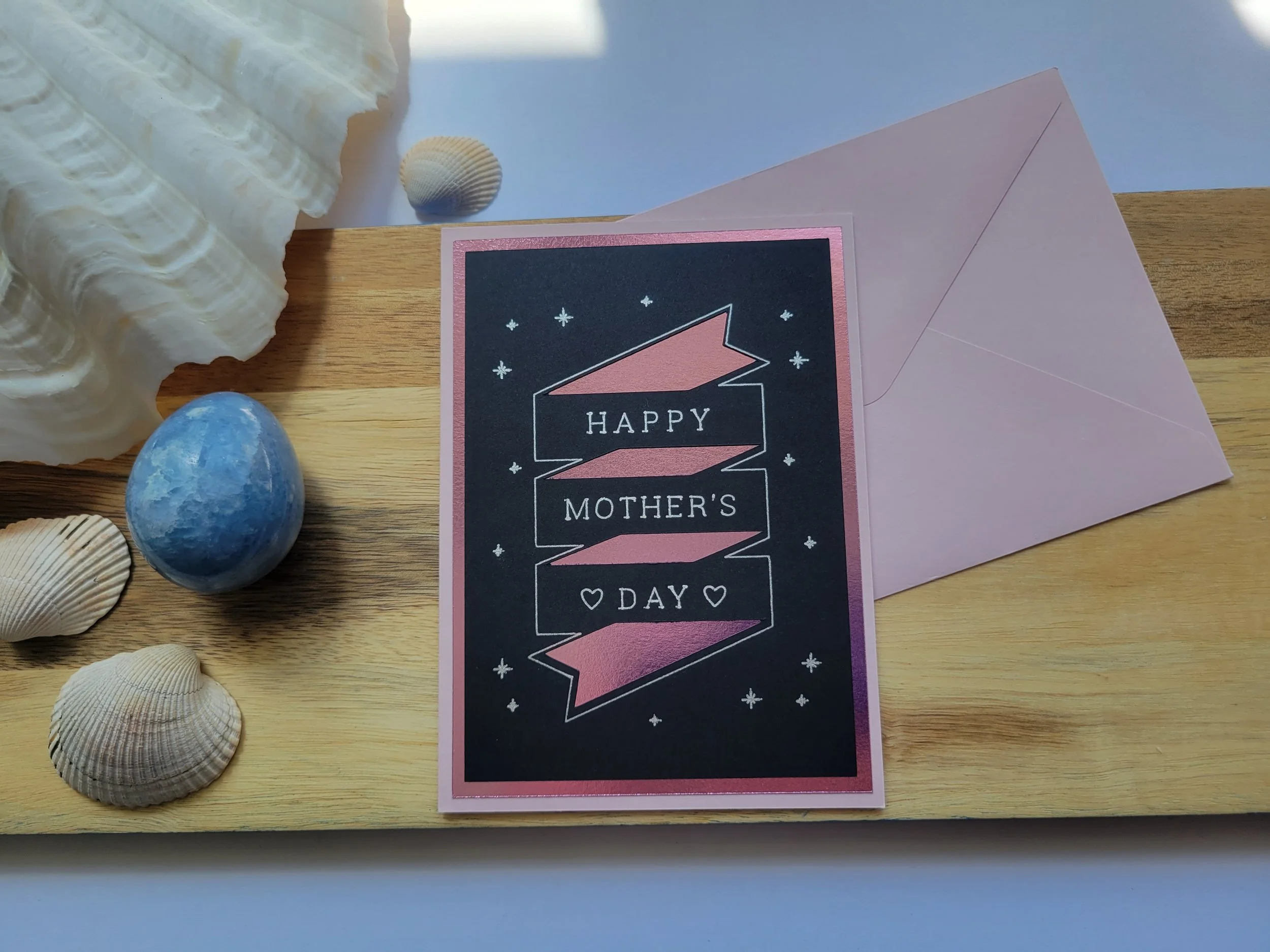 ink Greeting card with pink metallic banner design on black cardstock and the message “Happy Mother’s Day” placed on a wooden surface with seashells and a matching envelope.