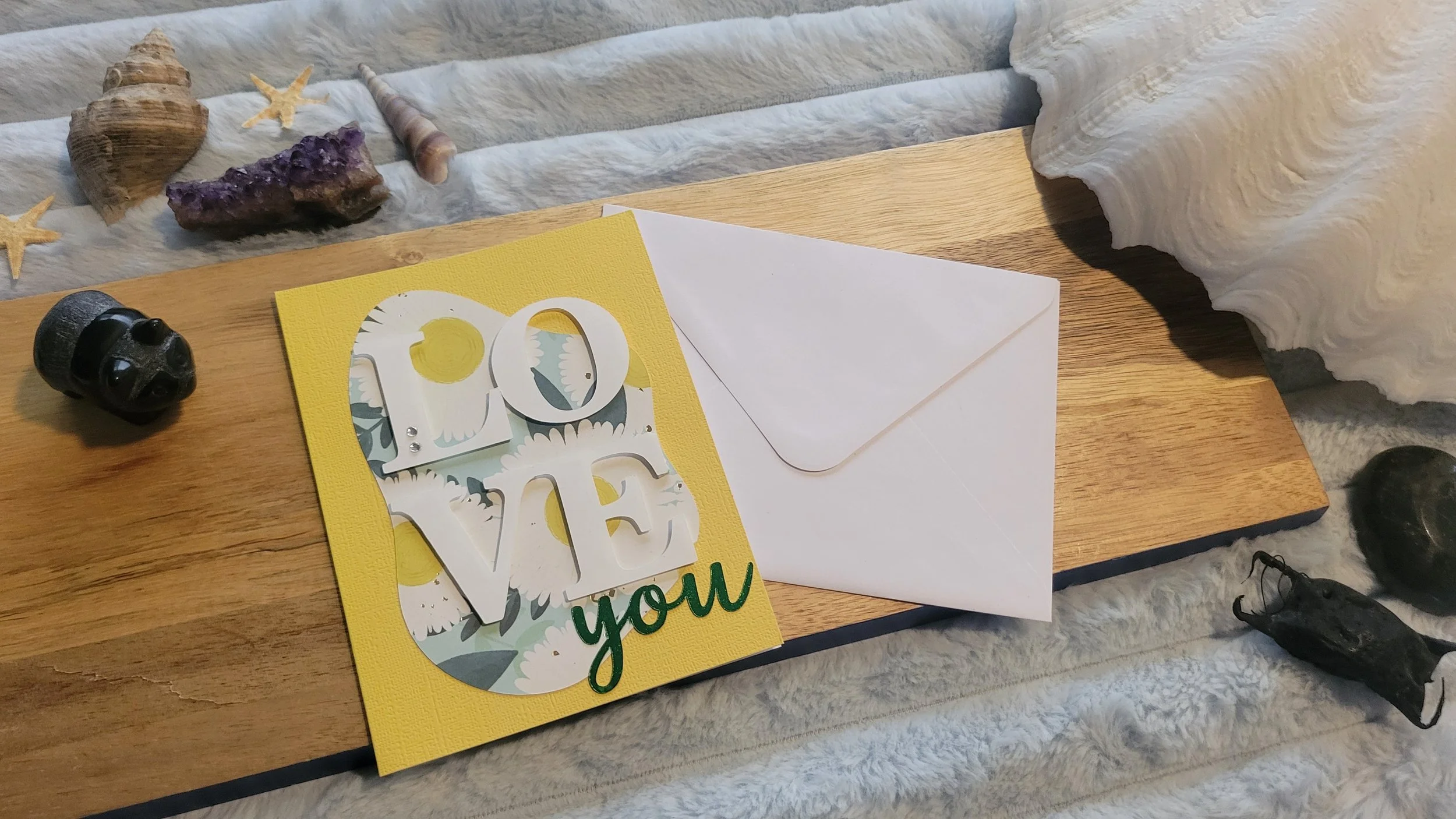 Yellow handmade card with a sunflower-patterned abstract shape, layered white “LOVE” lettering, and shiny green script “you” at the bottom