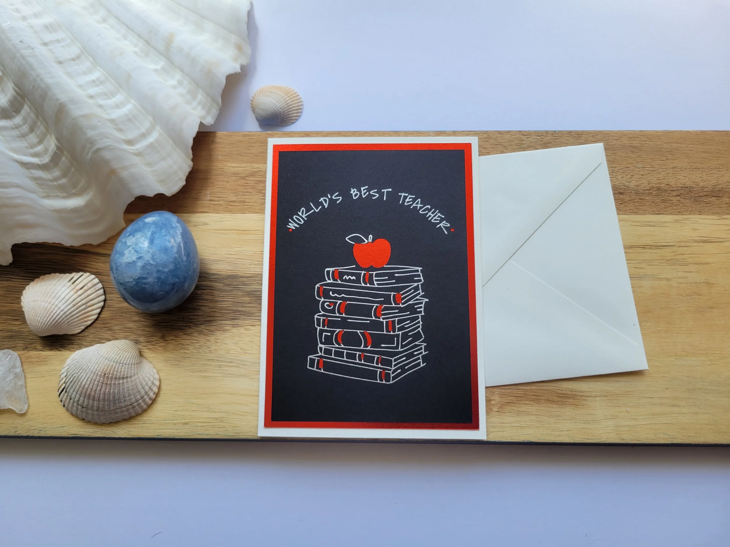 Handmade greeting card with a black background, red foil border, and illustration of stacked books with an apple, reading “World’s Best Teacher.”