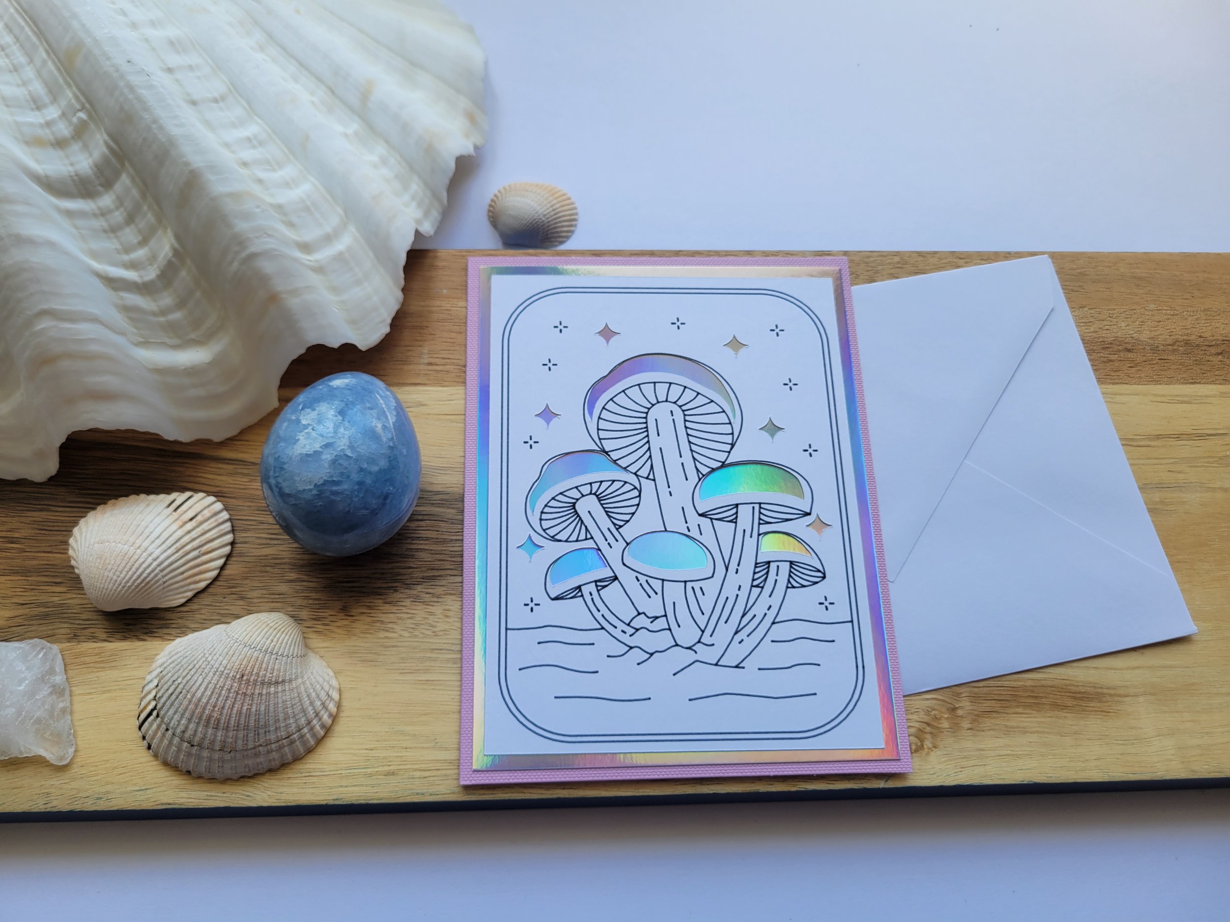 Minimalist mushroom illustration card with color-shifting foil details and a soft holographic border, displayed with a white envelope on a wooden board.