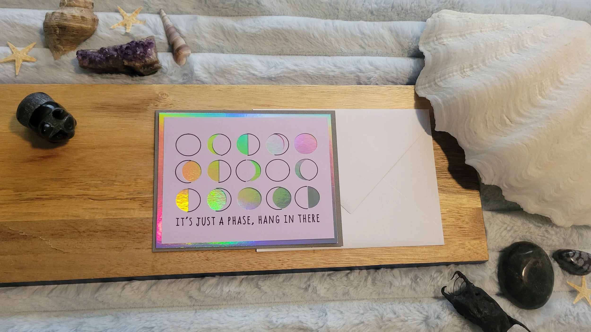 Handmade greeting card featuring moon phases in a grid with iridescent foil accents and the phrase “It’s just a phase, hang in there” on a holographic border.