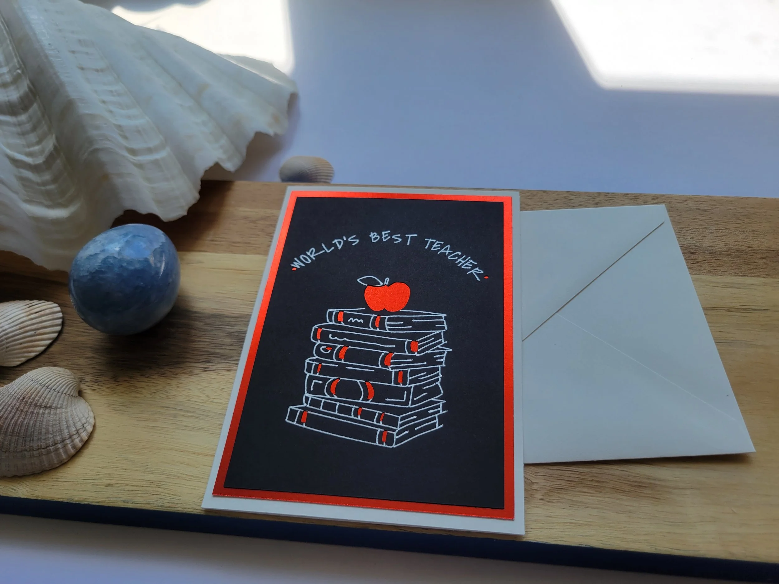 Handmade greeting card with a black background, red foil border, and illustration of stacked books with an apple, reading “World’s Best Teacher.”