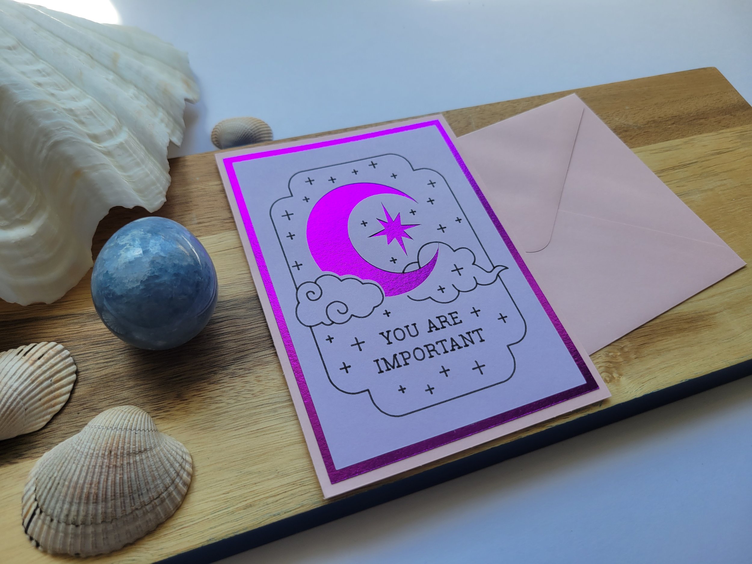 Pink Greeting card with purple metallic crescent moon and star design and the message “You Are Important,” placed on a wooden surface with seashells and a matching envelope.