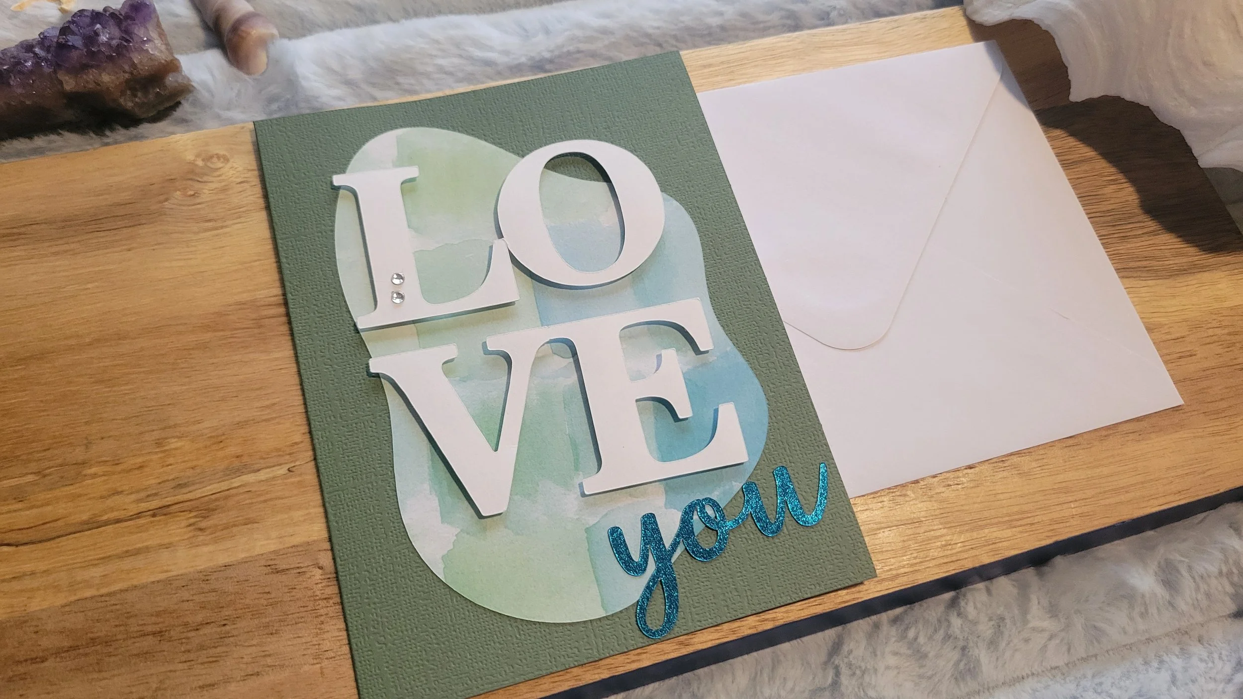 Green handmade card with a blue-green watercolor abstract shape, layered white “LOVE” lettering, and shiny blue script “you” at the bottom.