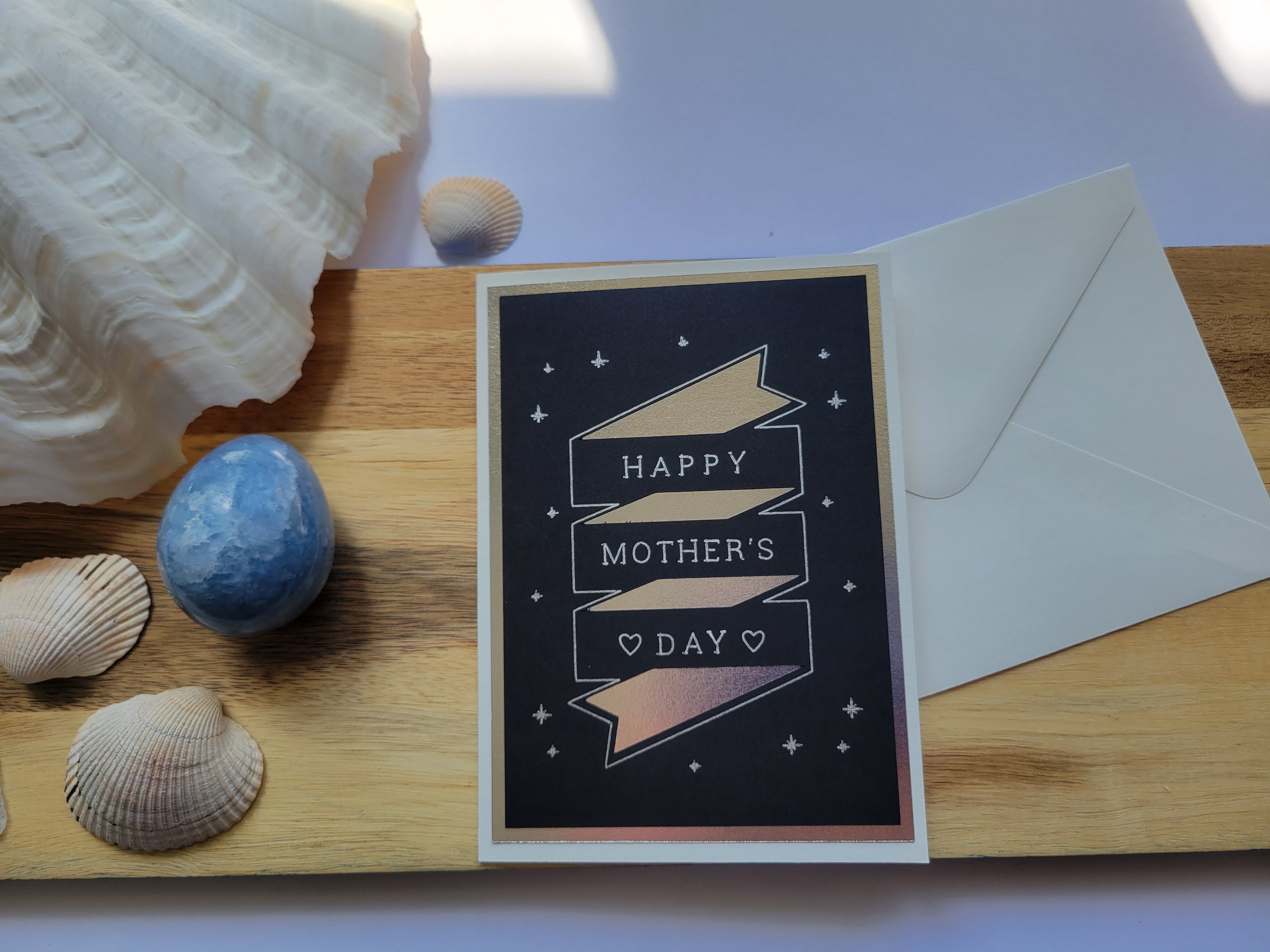 White Greeting card with silver metallic banner design on black cardstock and the message “Happy Mother’s Day” placed on a wooden surface with seashells and a matching envelope.