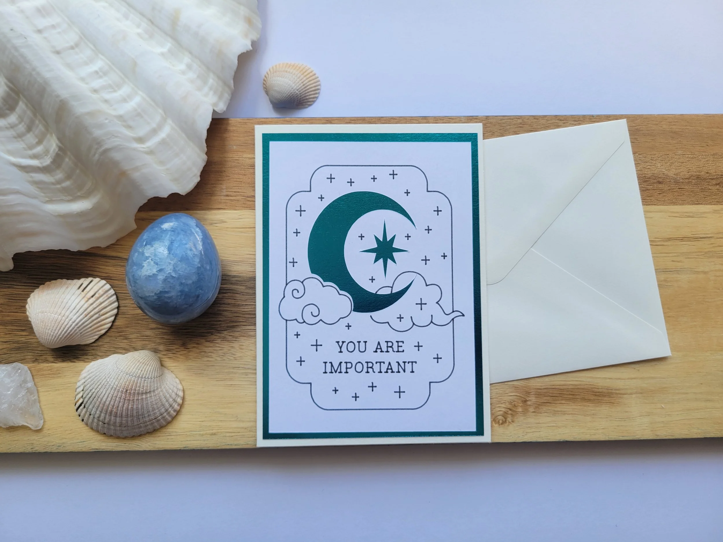 White Greeting card with teal metallic crescent moon and star design and the message “You Are Important,” placed on a wooden surface with seashells and a matching envelope.