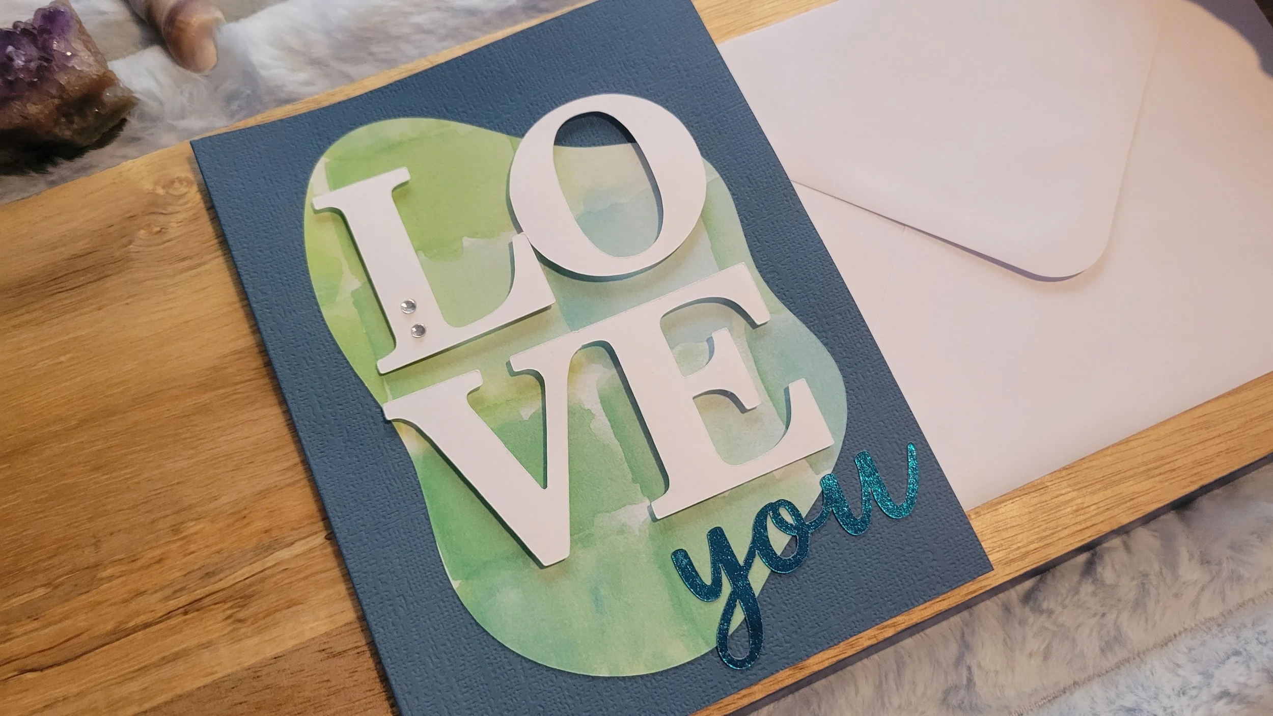 Navy handmade card with a blue-green watercolor abstract shape, layered white “LOVE” lettering, and shiny blue script “you” at the bottom.