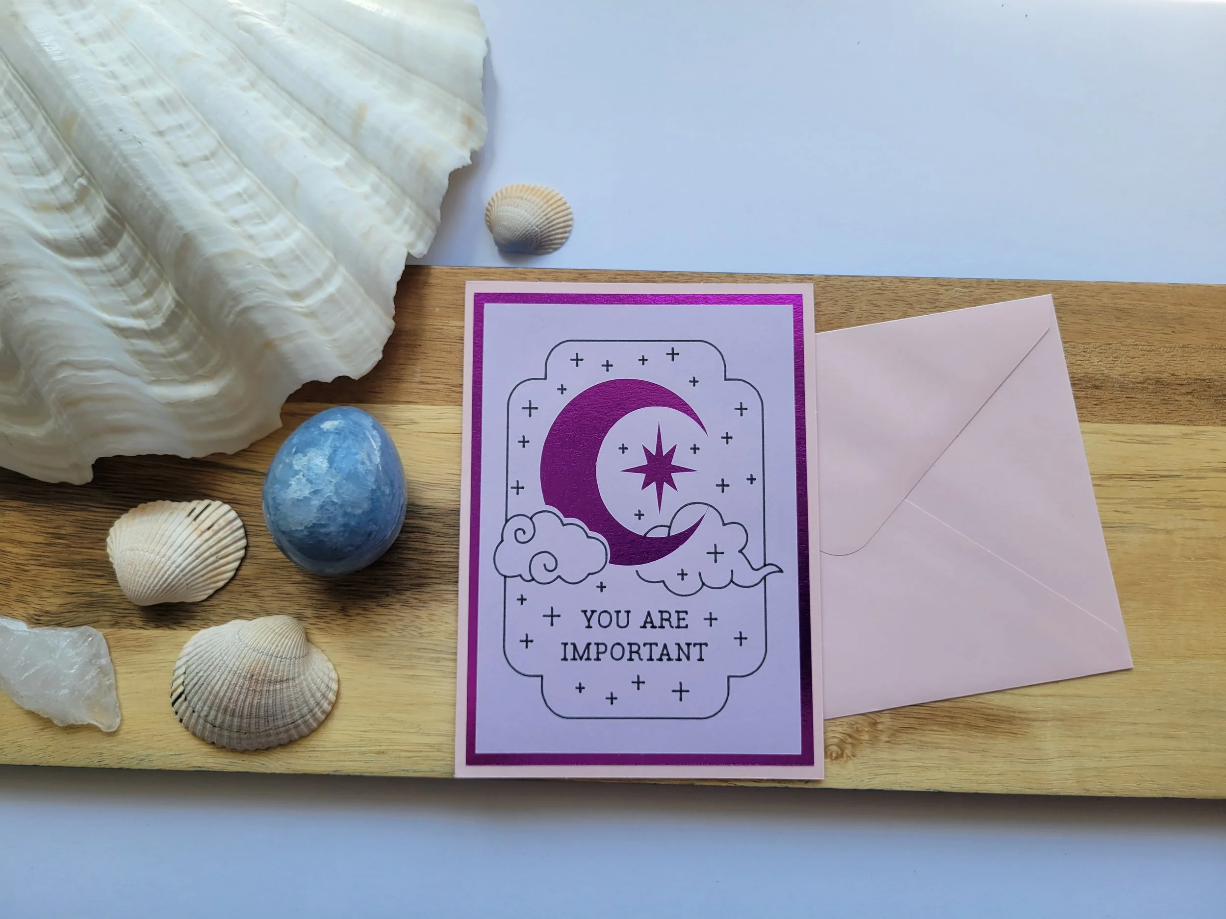 Pink Greeting card with purple metallic crescent moon and star design and the message “You Are Important,” placed on a wooden surface with seashells and a matching envelope.