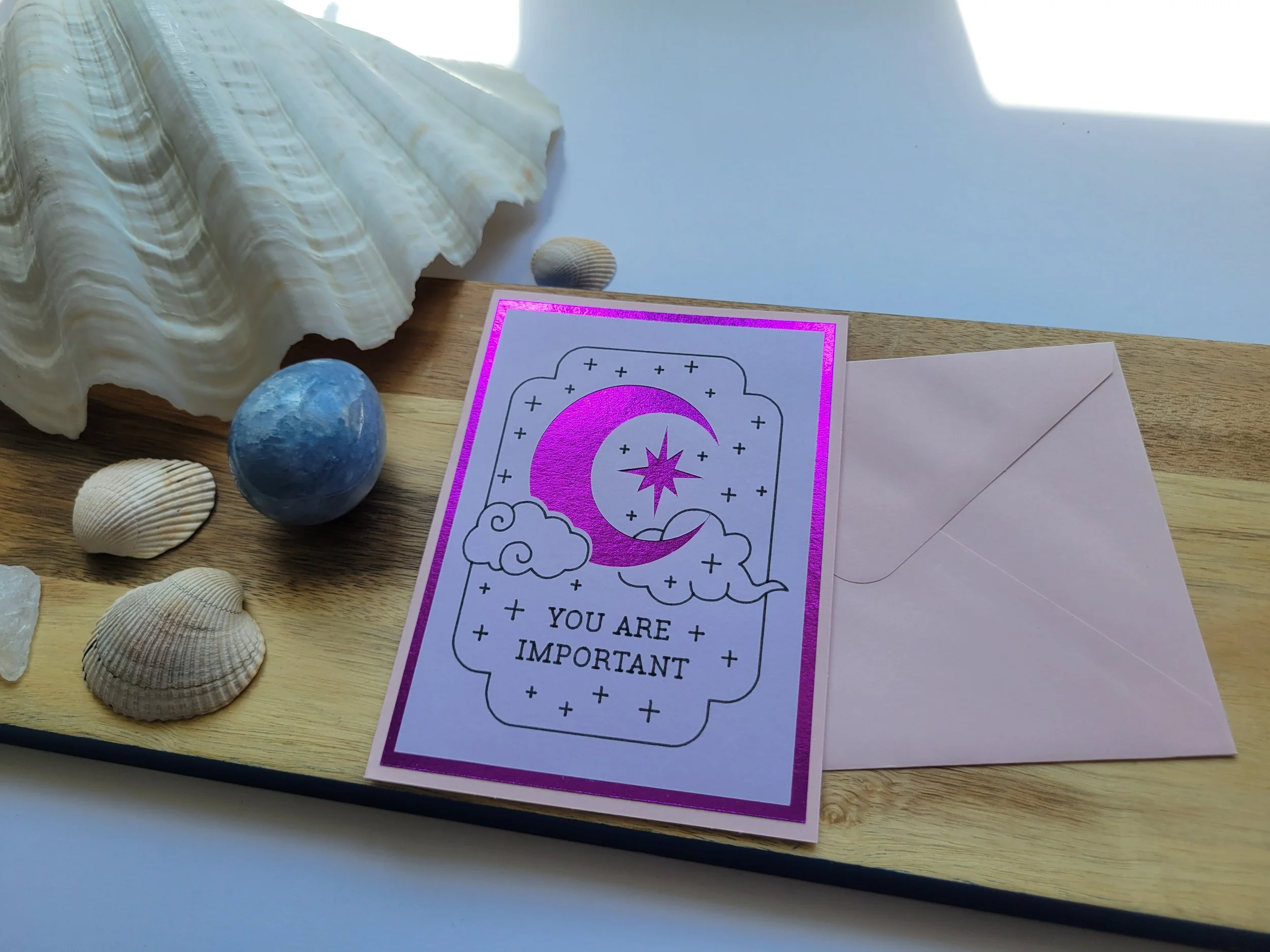 Pink Greeting card with purple metallic crescent moon and star design and the message “You Are Important,” placed on a wooden surface with seashells and a matching envelope.