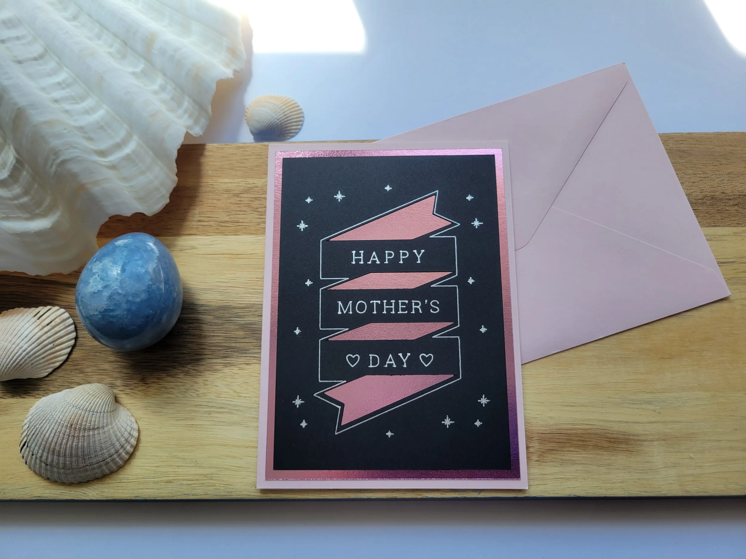 ink Greeting card with pink metallic banner design on black cardstock and the message “Happy Mother’s Day” placed on a wooden surface with seashells and a matching envelope.