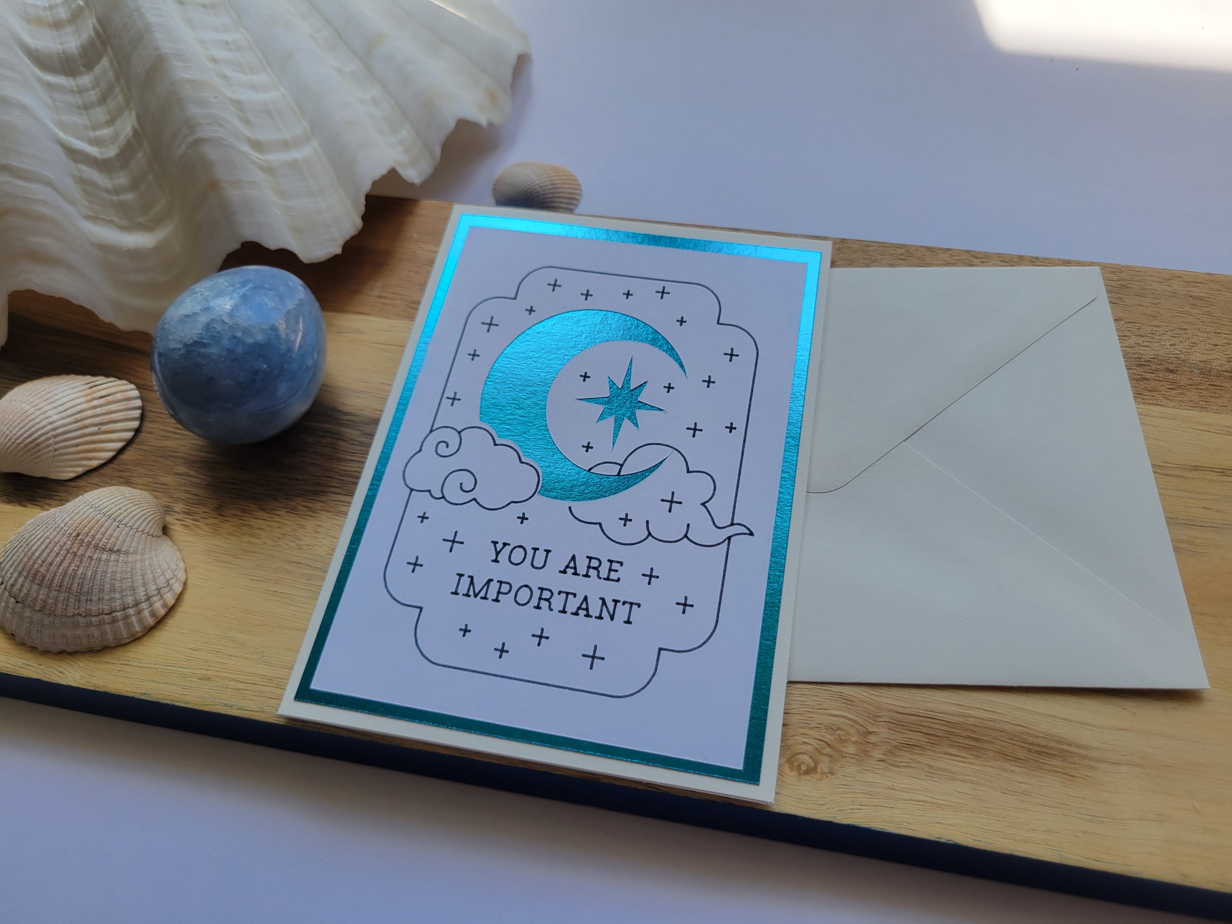 White Greeting card with teal metallic crescent moon and star design and the message “You Are Important,” placed on a wooden surface with seashells and a matching envelope.