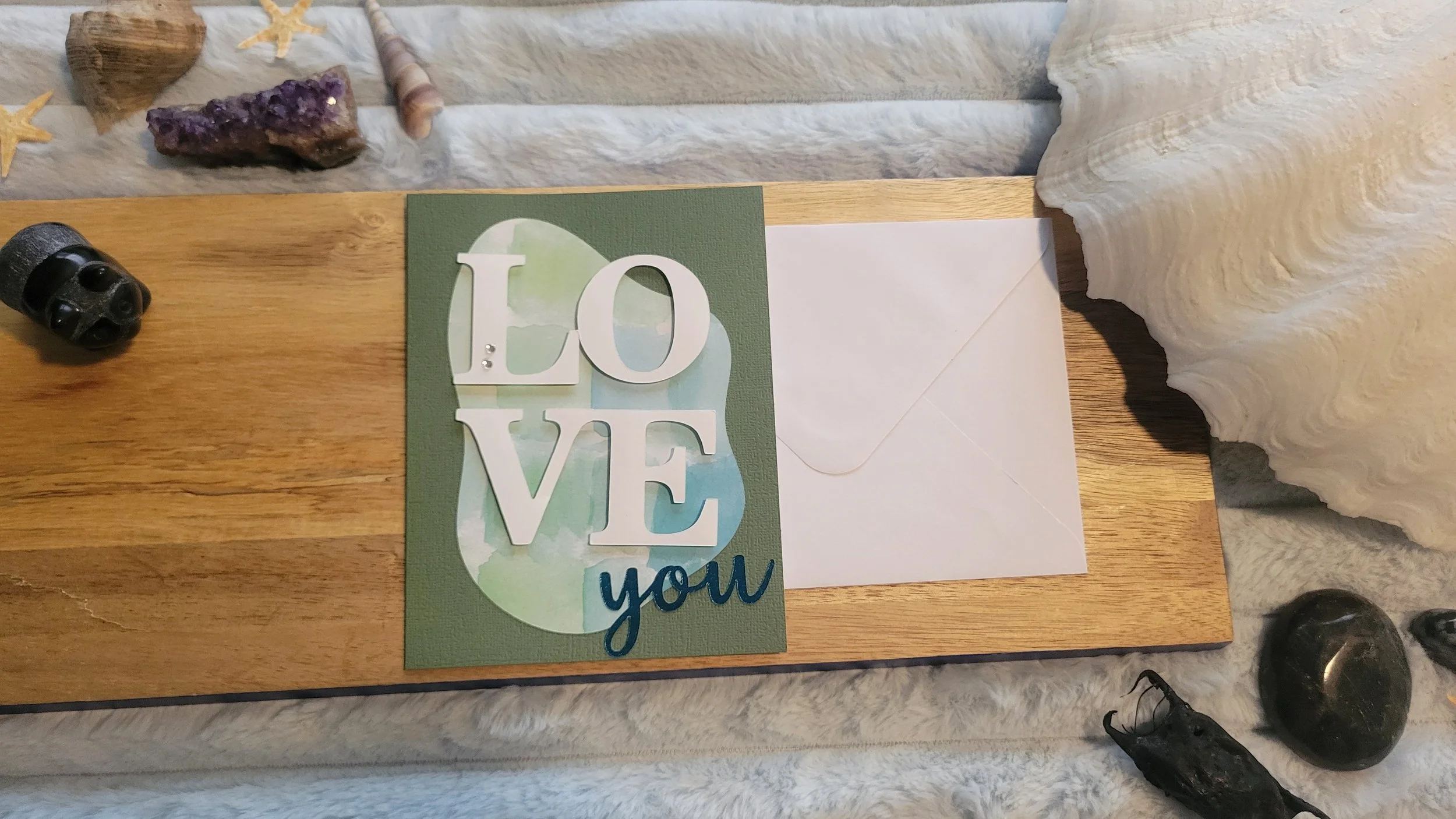 Green handmade card with a blue-green watercolor abstract shape, layered white “LOVE” lettering, and shiny blue script “you” at the bottom.