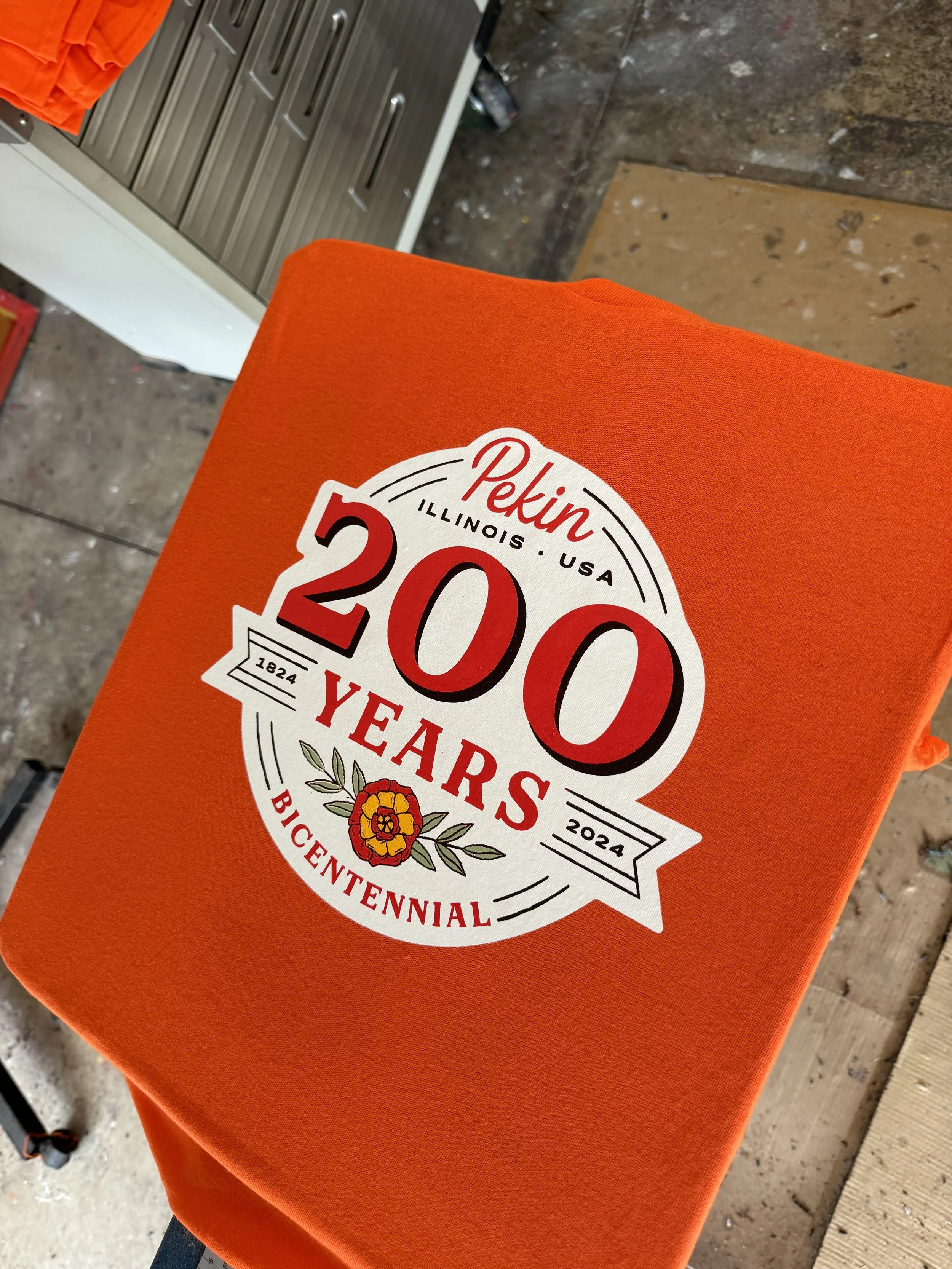 Orange T-shirt with a printed 200th anniversary logo for Pekin, Illinois, featuring a flower, the years 1824 and 2024, and the words '200 Years Bicentennial'.