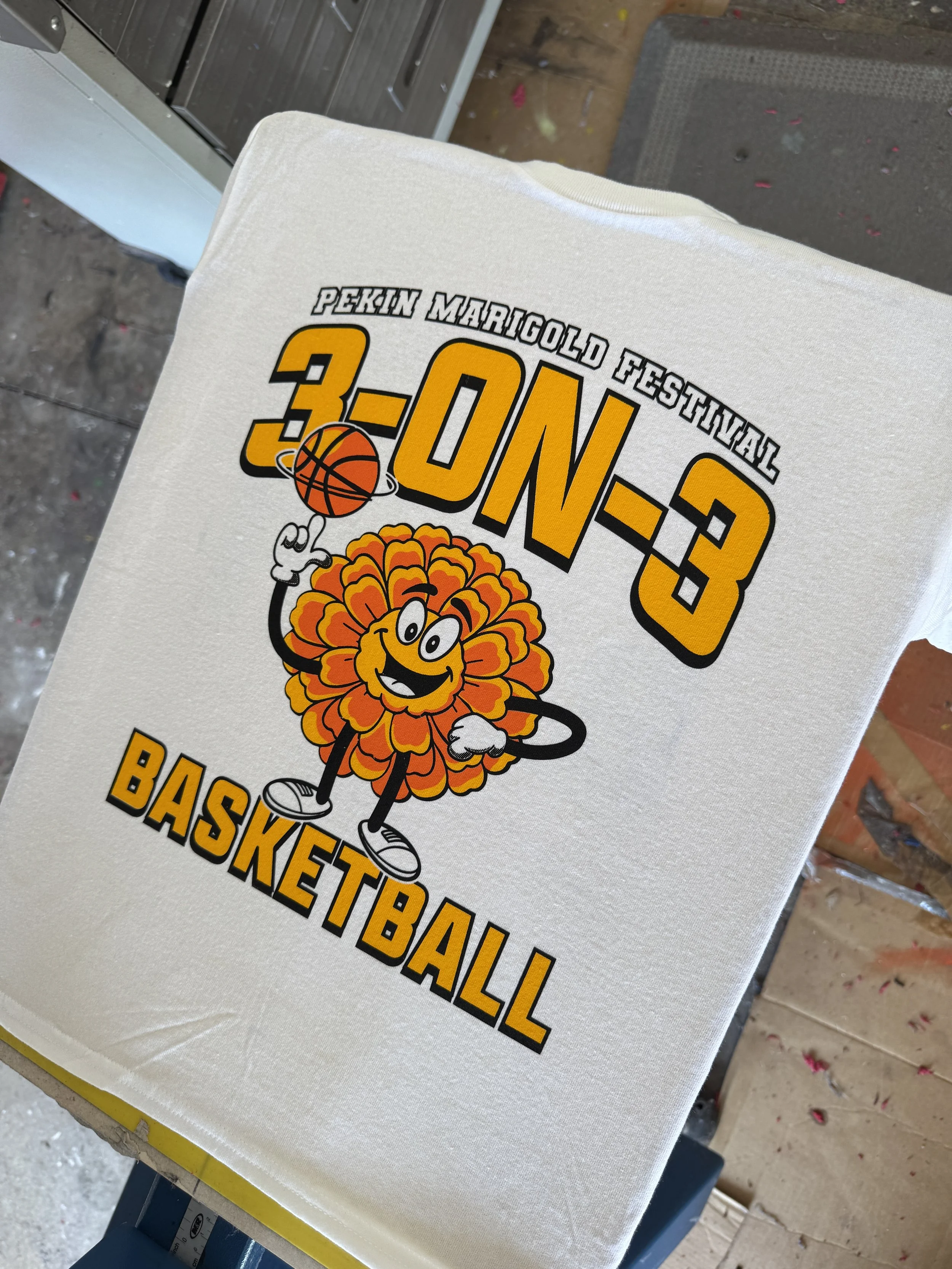 White T-shirt with a cartoon marigold flower character dribbling a basketball, promoting the Pekin Marigold Festival 3-on-3 basketball tournament.